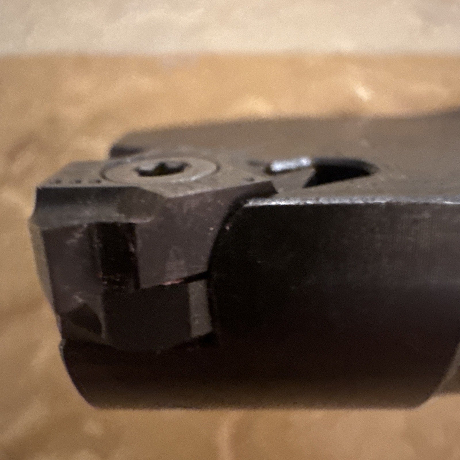 Close-up of a dark metal cutting tool tip with a curved edge and a screw-securing mechanism, set against a blurred beige background.
