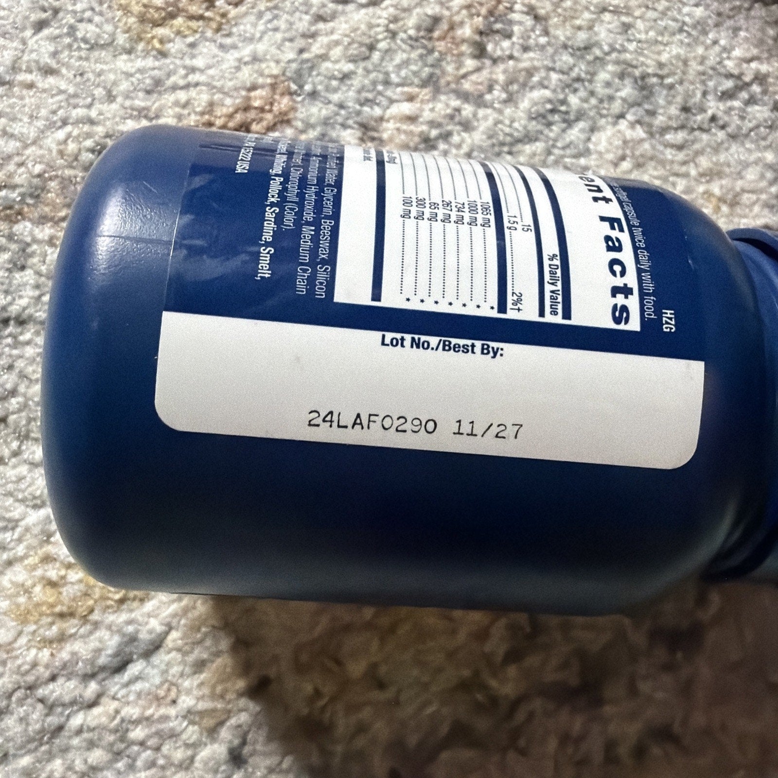 Close-up of a blue supplement bottle lying on a textured carpet, showing a white label with a lot number and expiration date printed as "24LAF0290 11/27" next to partial nutritional facts.