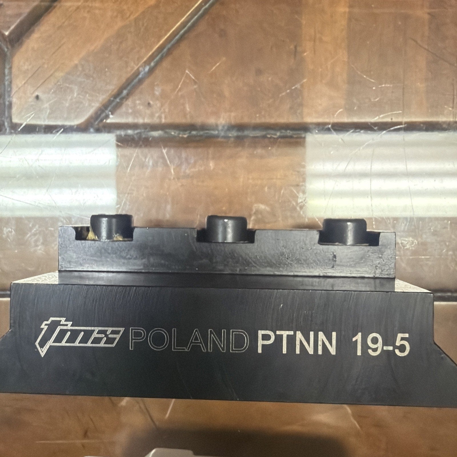 Black metal tool block engraved with "TMG Poland PTNN 19-5," featuring three cylindrical protrusions, resting on a reflective wooden surface.