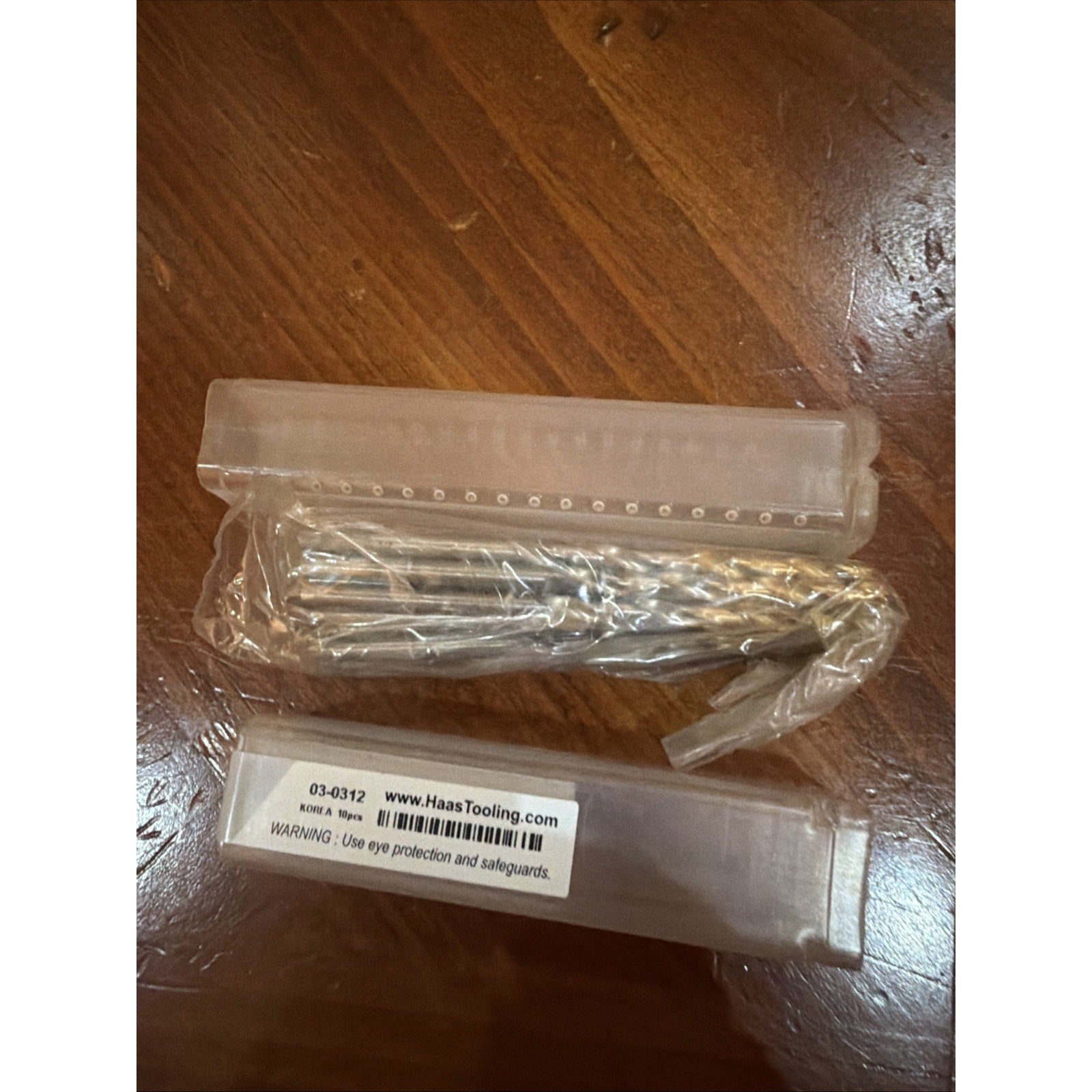 Several metal tool bits wrapped in clear plastic, resting next to a transparent plastic storage case with a white label displaying a web address and safety warning, all placed on a wooden surface.