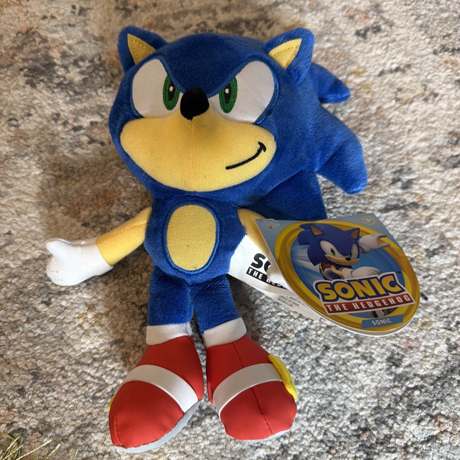 Plush toy of Sonic the Hedgehog with blue fur, green eyes, white gloves, red shoes, and an attached circular tag featuring Sonic artwork and logo, resting on a textured carpet.