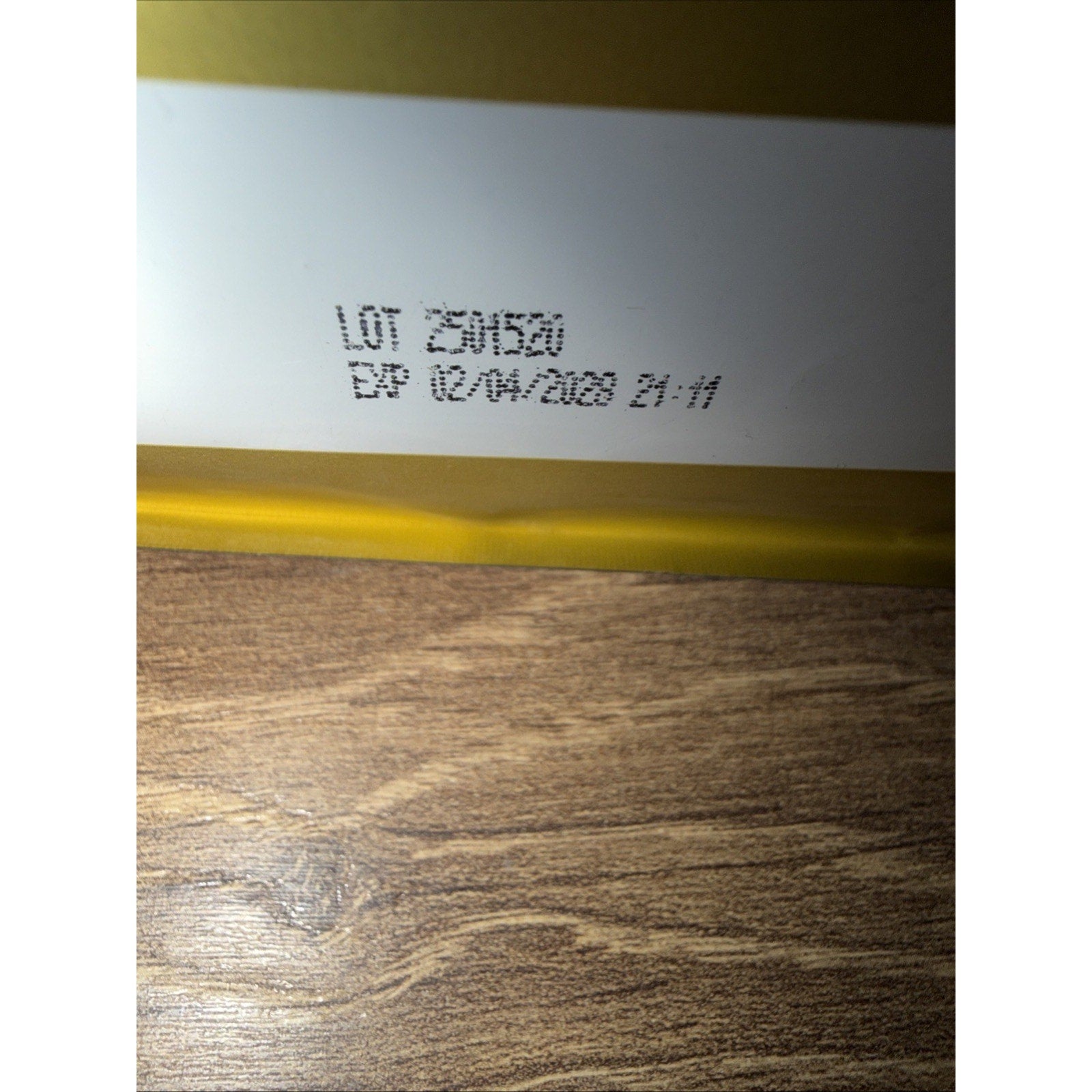 Close-up of a product package corner showing a lot number and expiration date printed in black ink on a white and yellow background over a wooden surface.