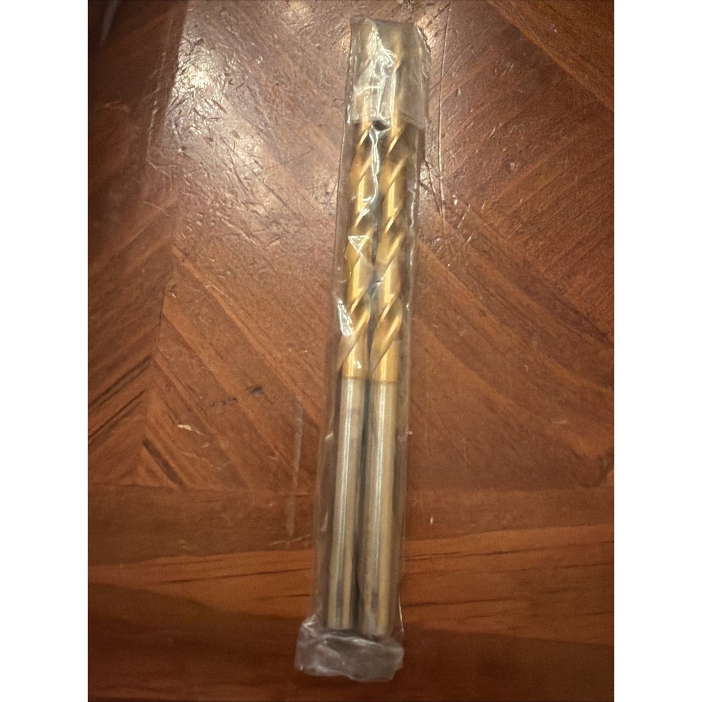 Two metal twist drill bits with gold-colored spiral flutes inside clear plastic packaging, placed on a wooden surface with a parquet pattern.