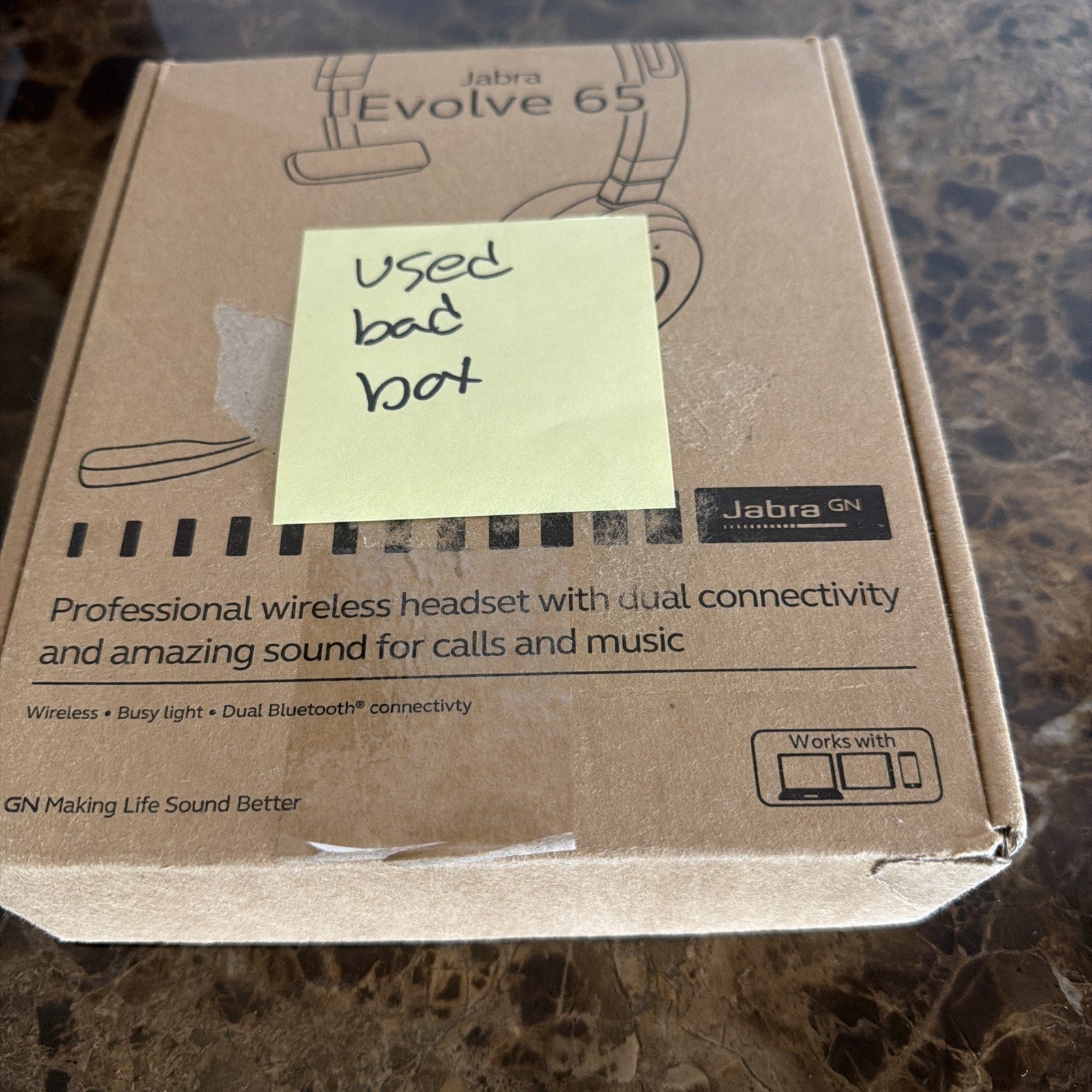 Brown cardboard box packaging for a Jabra Evolve 65 wireless headset with printed product features, a headset illustration, and a yellow sticky note with handwritten text placed on top.