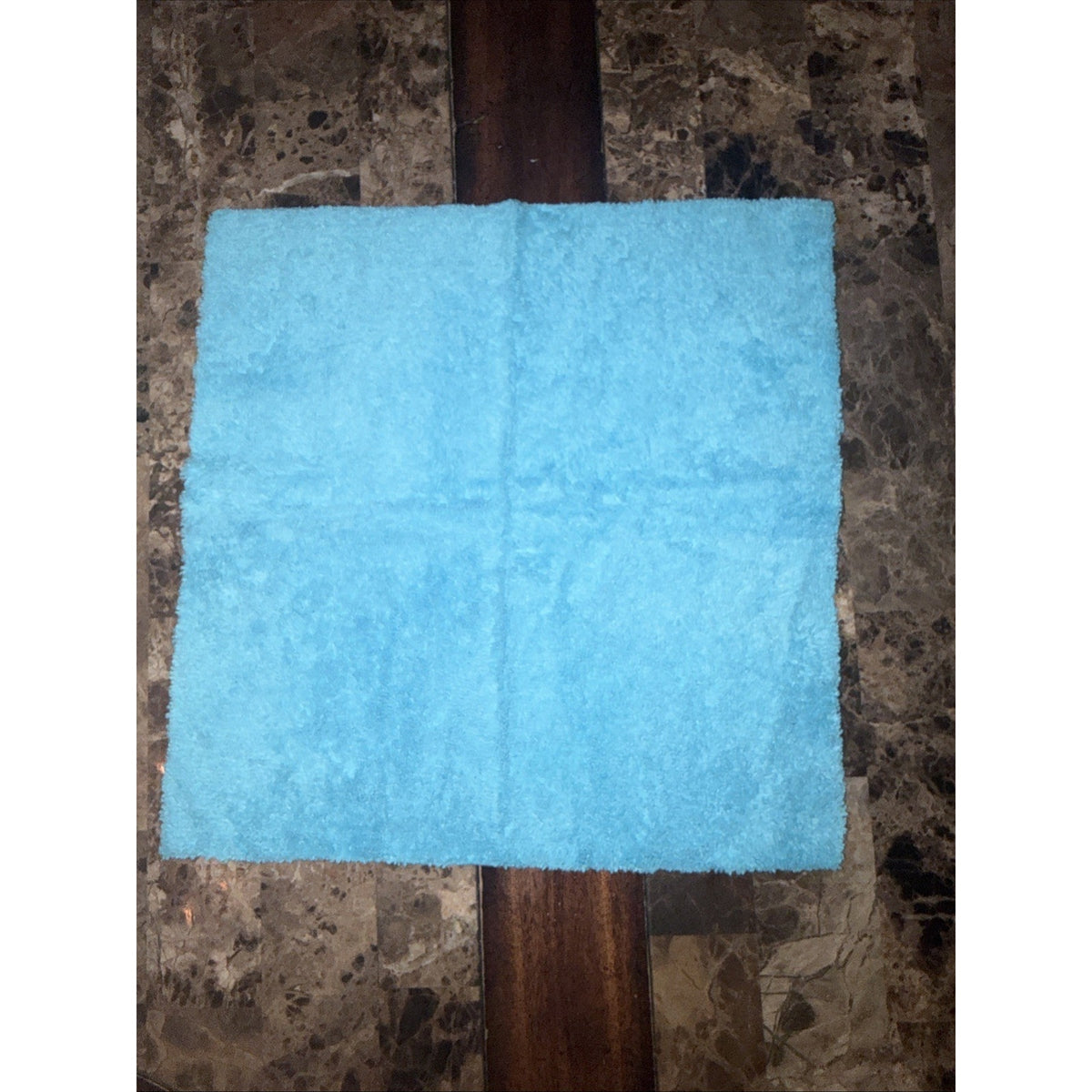 Soft light blue microfiber cleaning cloth laid flat on a dark marble surface with a wooden strip running vertically beneath it.