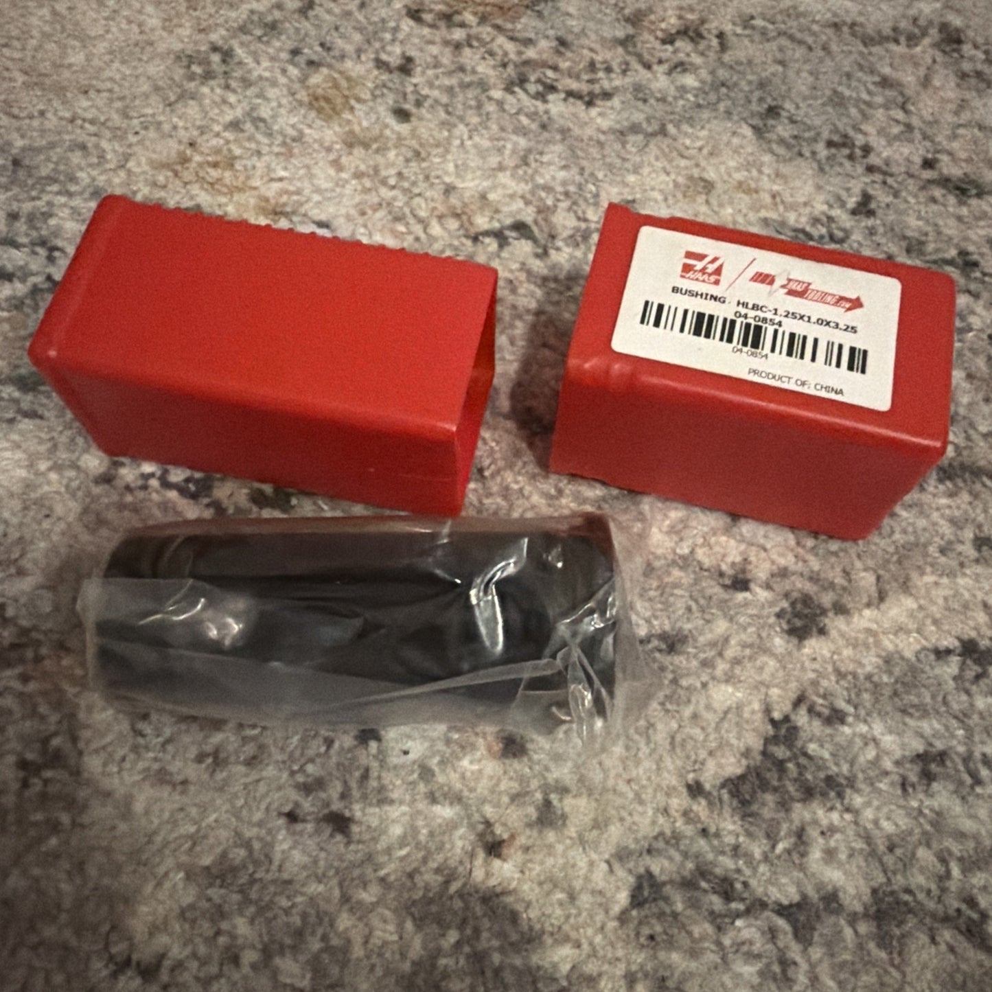 Two red rectangular plastic containers, one with a white label featuring a barcode and text, alongside a black cylindrical tool wrapped in clear plastic, all on a textured fabric surface.