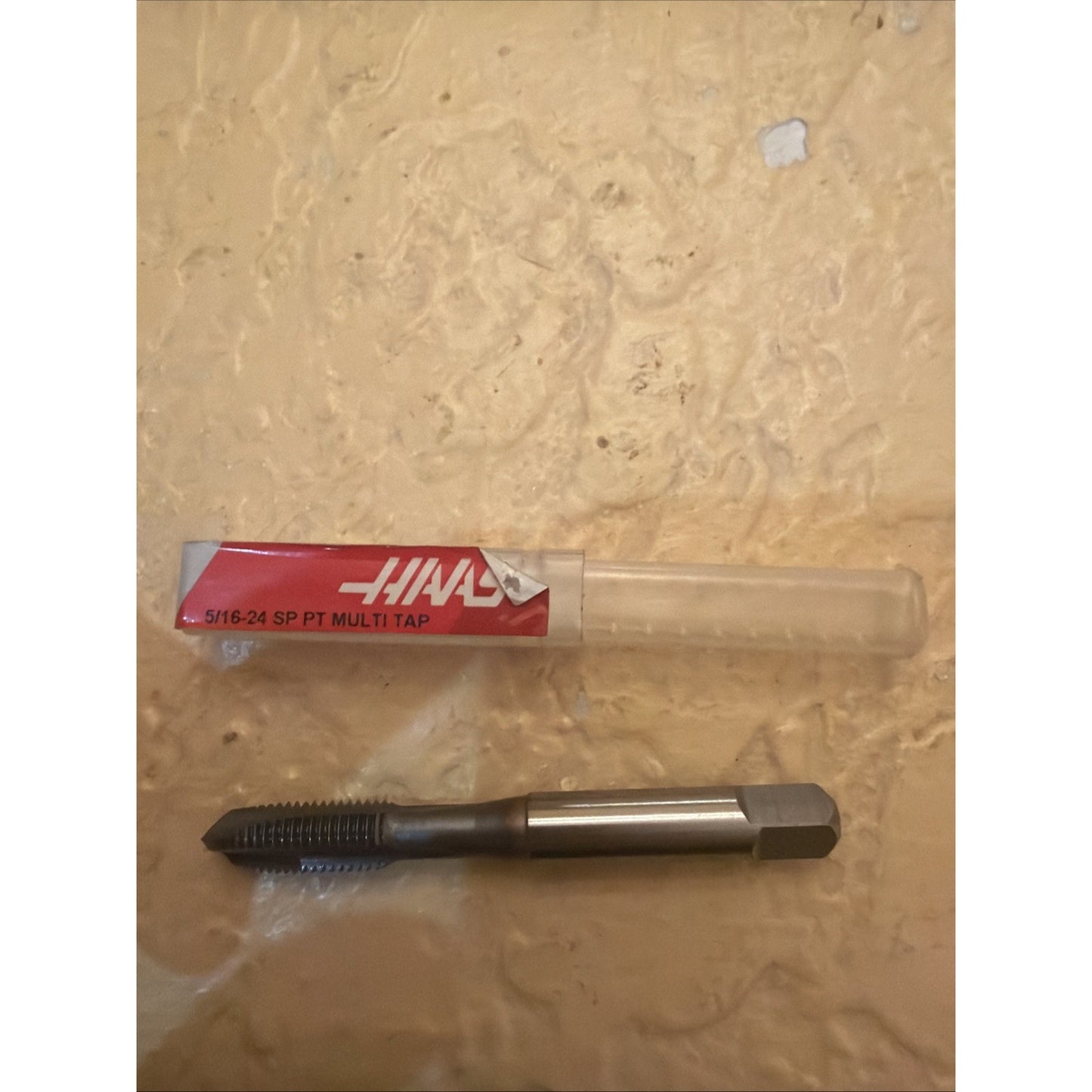 Metal threading tap with sharp threads and a hexagonal shank, displayed next to a clear plastic storage tube with a red and white label indicating size and type.