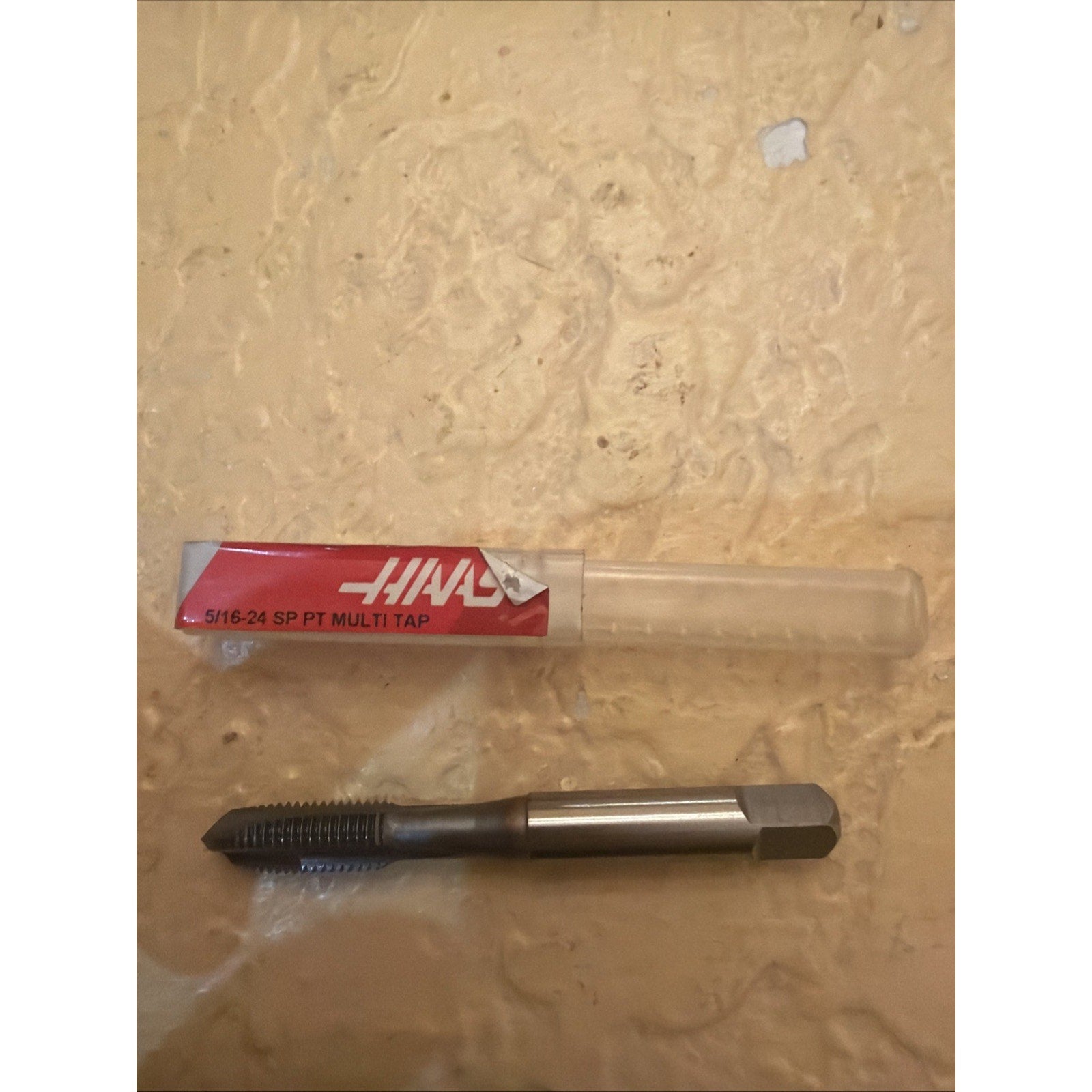 Metal threading tap with sharp threads and a hexagonal shank, displayed next to a clear plastic storage tube with a red and white label indicating size and type.