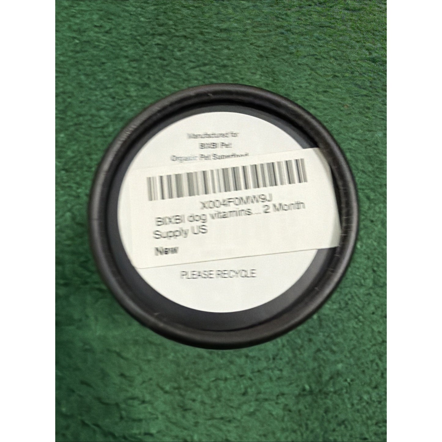 Top view of a black container lid with a white label showing product information for a two-month supply of dog vitamins, placed on a green textured surface.