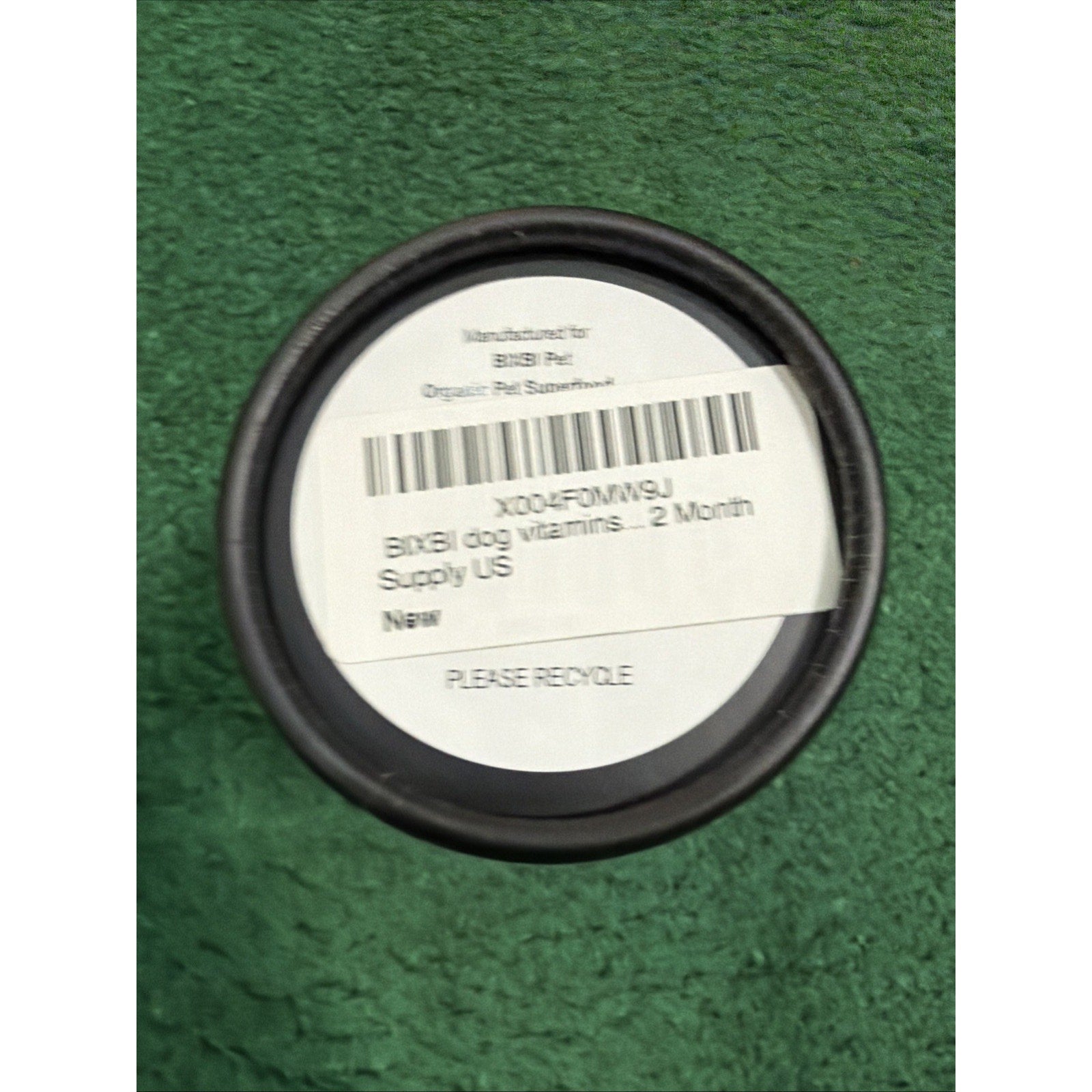 Top view of a black container lid with a white label showing product information for a two-month supply of dog vitamins, placed on a green textured surface.