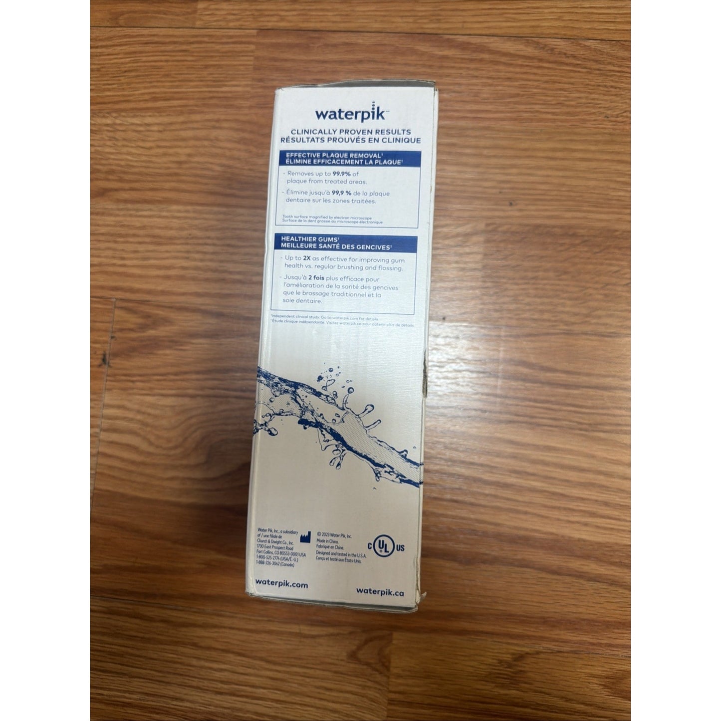 White rectangular product box for a Waterpik device, showing clinical results for plaque removal and gum health claims, with blue text and a water splash graphic on a wooden floor background.