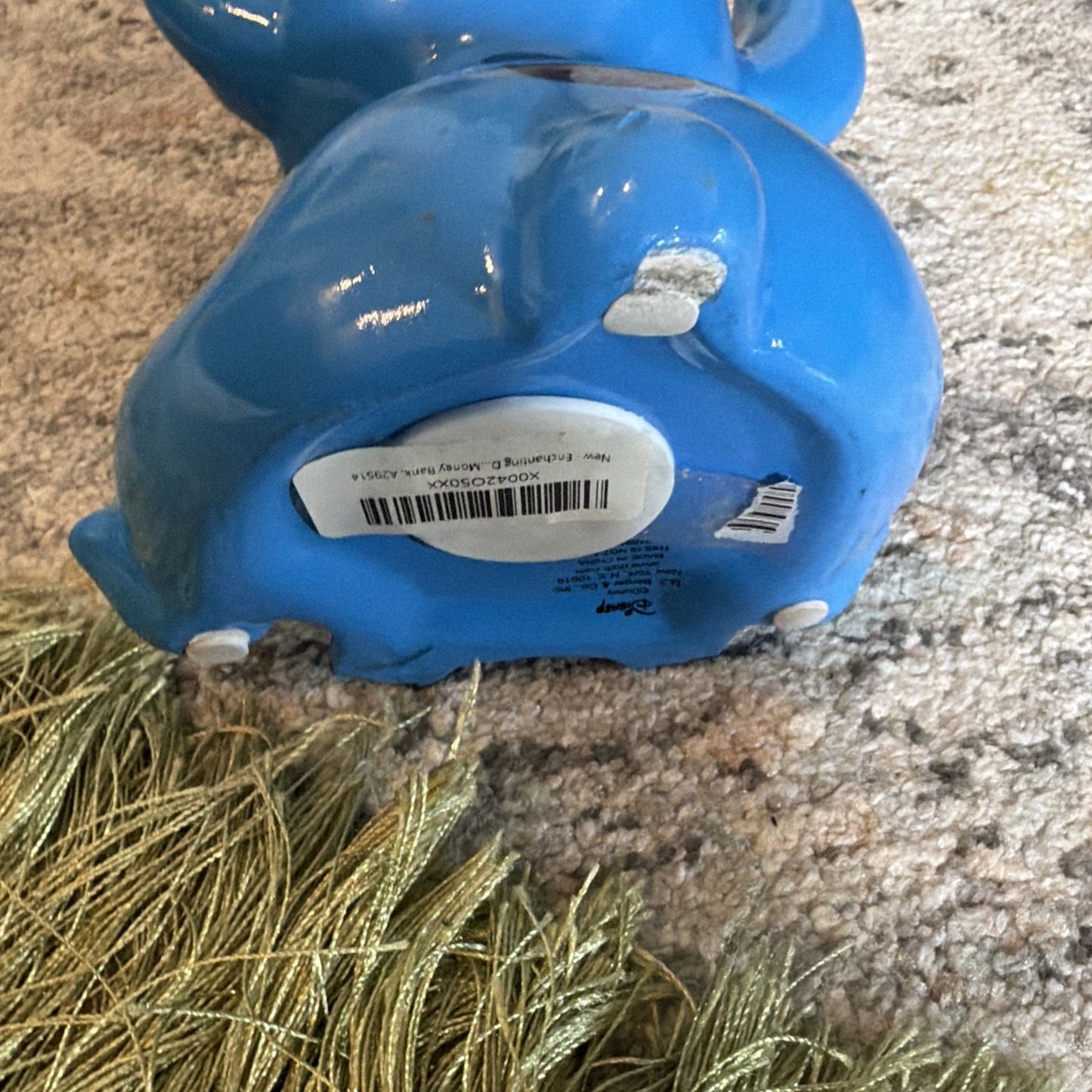Glossy blue ceramic figurine lying on its back on a textured carpet with a green tassel accessory nearby, showing the base with stickers and small white pads.
