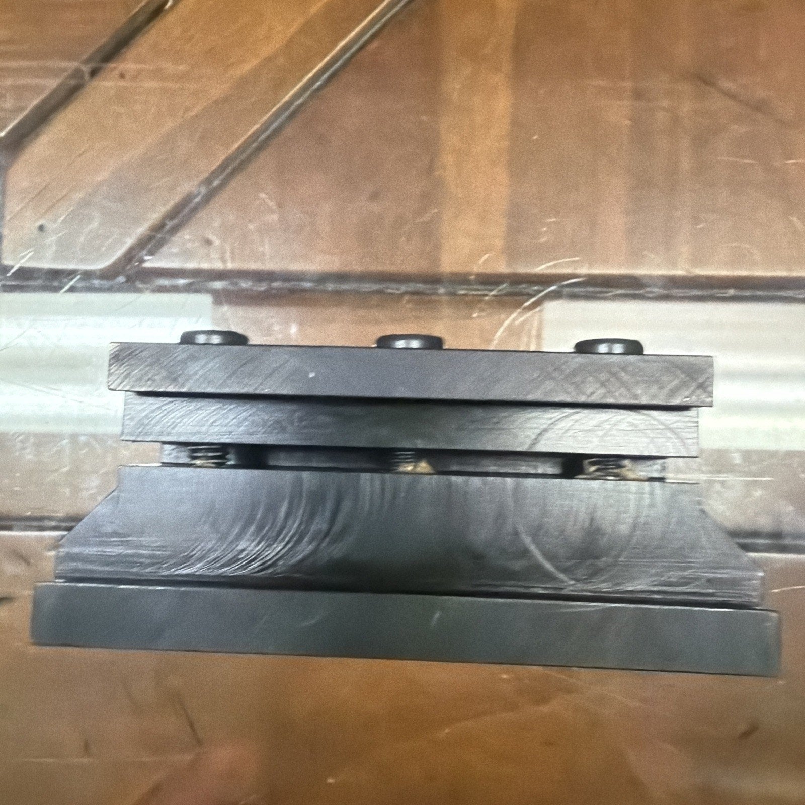 Black metal clamp component with three evenly spaced screws on top, resting on a transparent surface with visible reflections and scratches.