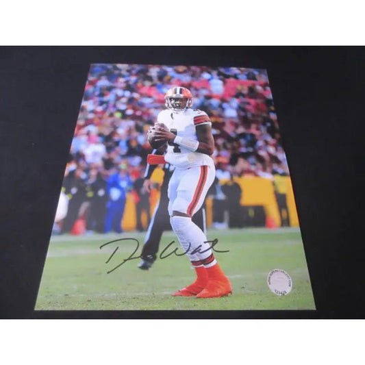 Printed photograph of a football player in white uniform with orange cleats mid-action on the field, featuring a visible autograph and a small authentication sticker in the corner.