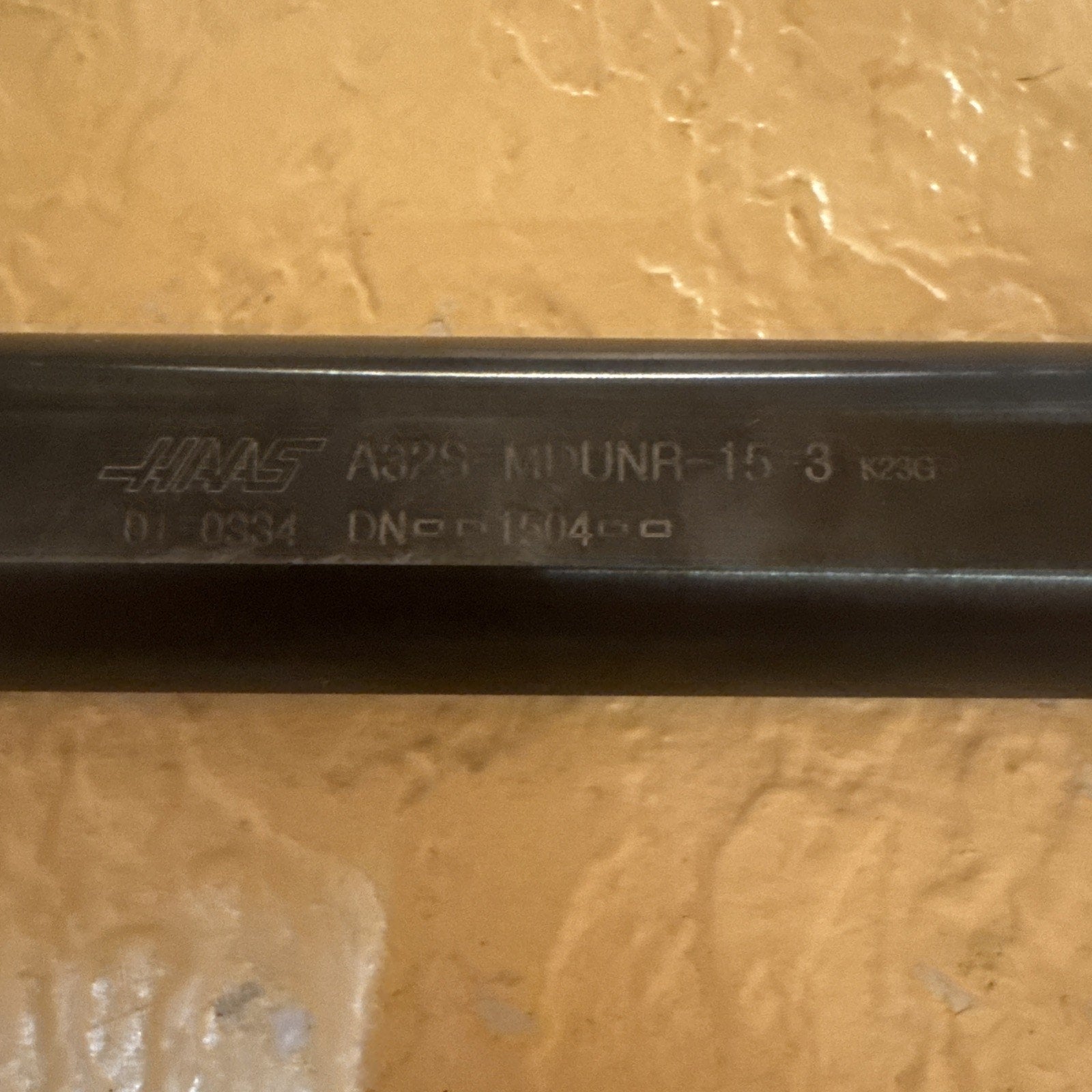 Close-up of a dark metal cylindrical tool shaft engraved with the Haas logo, model number A32S-MDUNR-15-3, and additional alphanumeric codes, set against a textured beige wall.