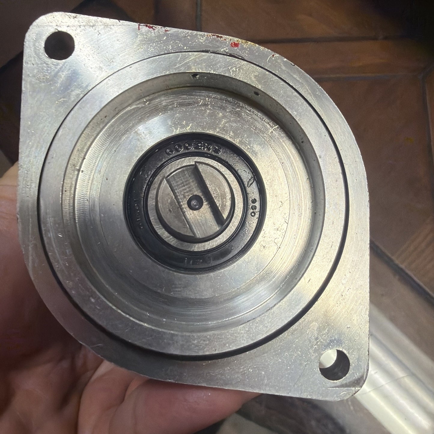 Hand holding a circular metal mechanical part with concentric grooves and a central black ring, featuring two mounting holes on a triangular flange, against a tiled floor background.