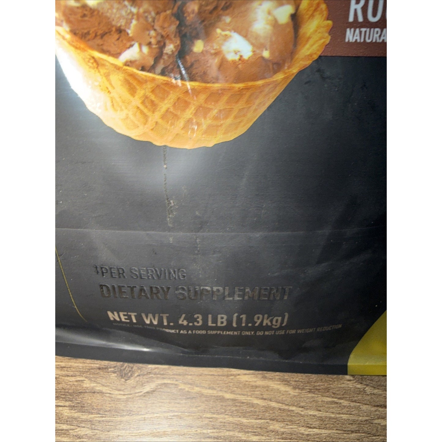 Close-up of a dietary supplement package showing a waffle cone filled with chocolate pieces and text indicating net weight of 4.3 pounds (1.9 kg) and serving information.