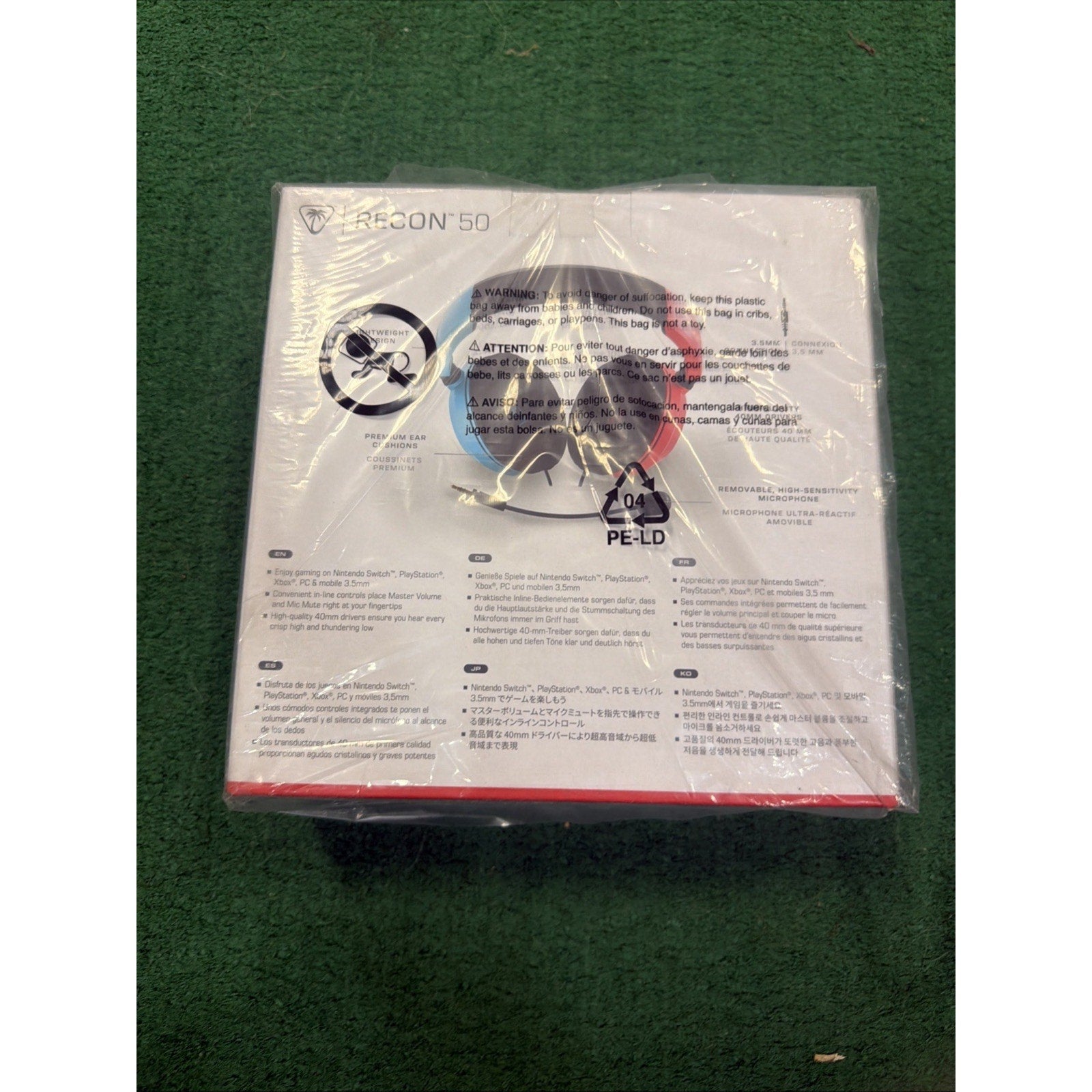 White product box for a Recon 50 gaming headset featuring blue and red ear cup illustrations, multilingual safety warnings, and a plastic wrap with a recycling symbol.