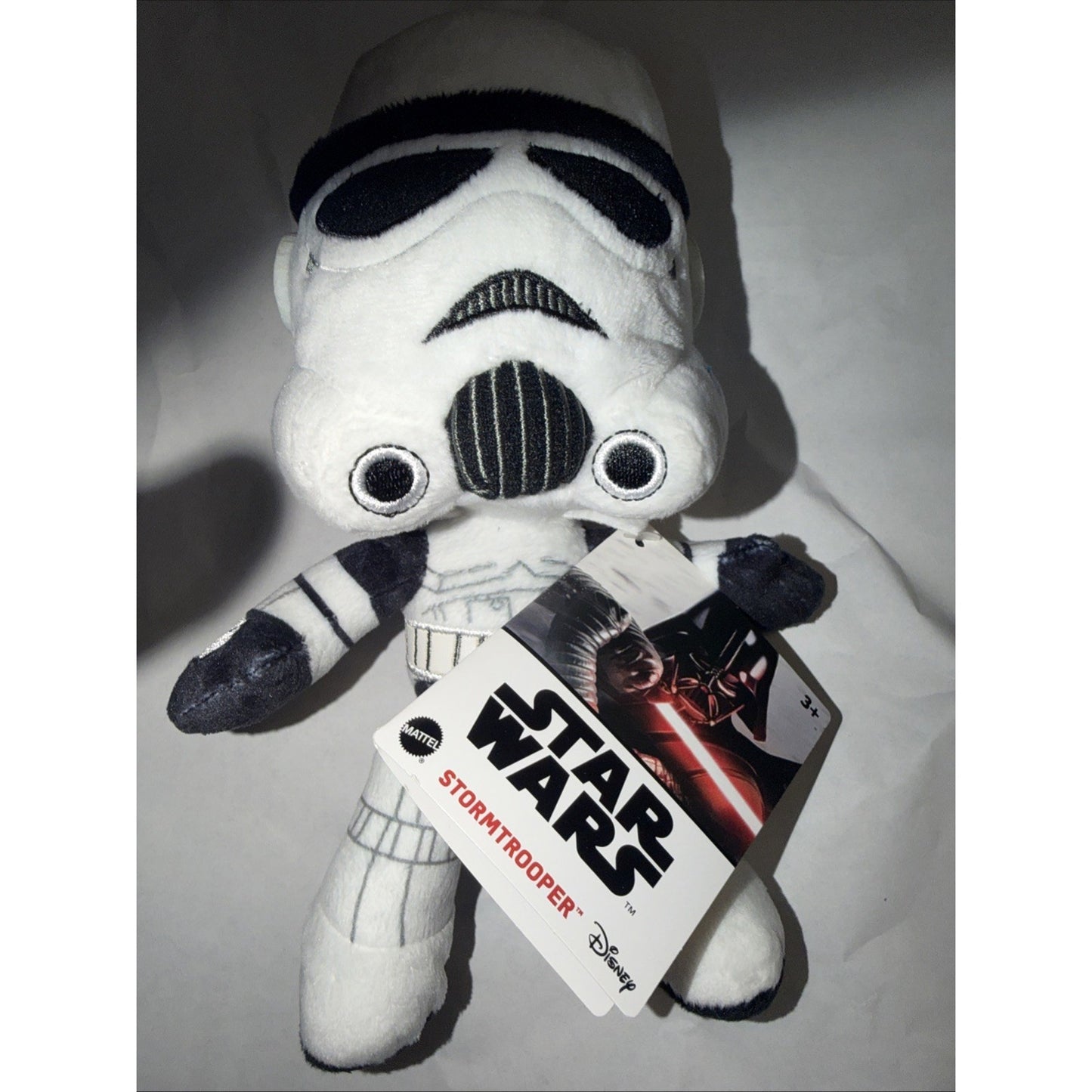 Plush toy of a Star Wars Stormtrooper wearing white armor with black details, holding a Star Wars and Disney-branded tag featuring Darth Vader on the cover.