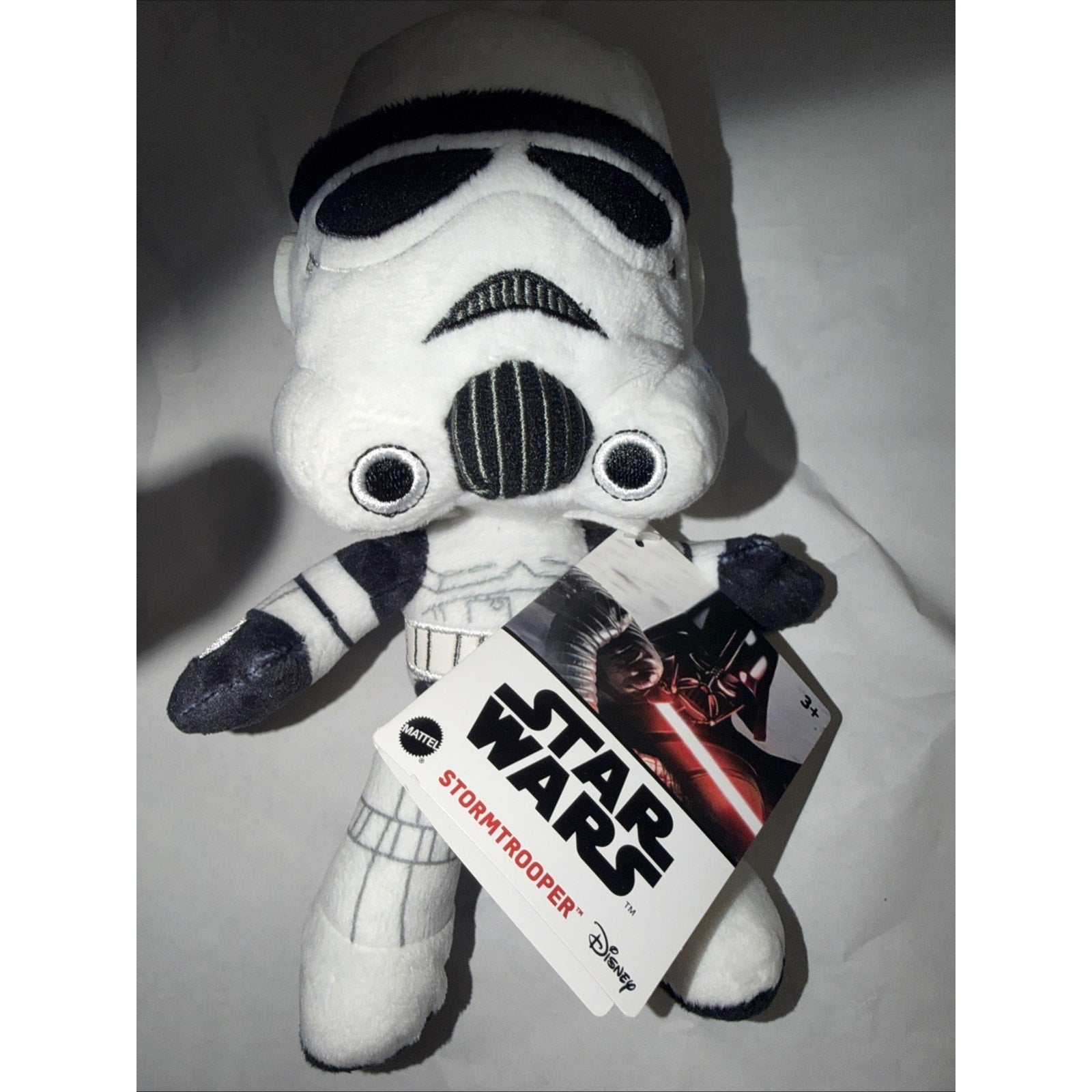 Plush toy of a Star Wars Stormtrooper wearing white armor with black details, holding a Star Wars and Disney-branded tag featuring Darth Vader on the cover.