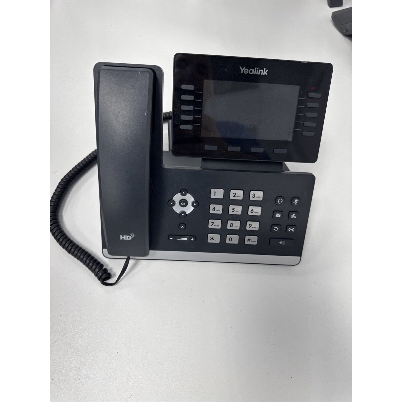 Black office desk phone with a digital display screen, numeric keypad, navigation buttons, and coiled handset cord placed on a white surface.