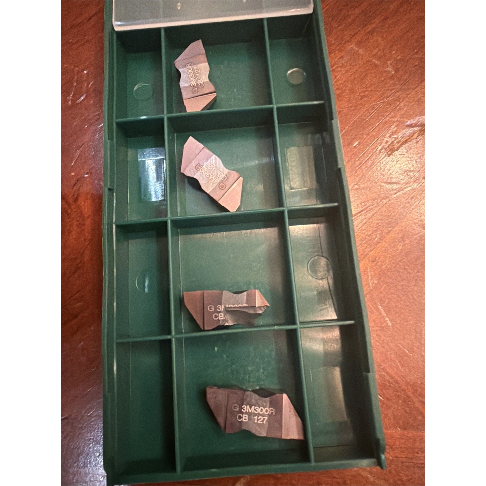 Green plastic organizer tray with multiple compartments, holding four small irregularly shaped metal cutting inserts with printed alphanumeric markings, placed on a wooden surface.