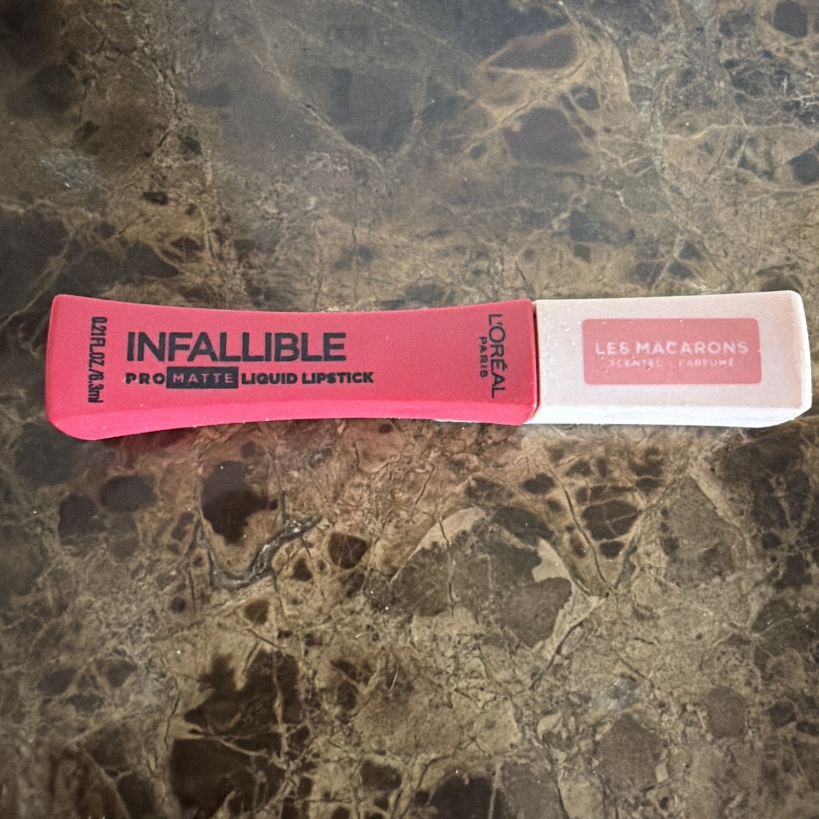 L'Oréal Paris Infallible Pro-Matte liquid lipstick in a vibrant coral pink shade with a white cap labeled "Les Macarons," placed on a dark marbled surface.