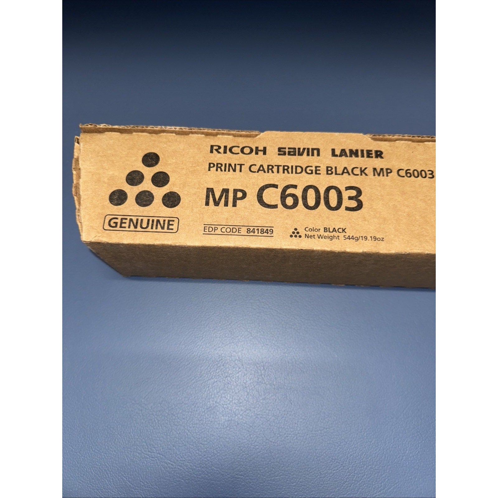 Brown cardboard packaging box labeled for a genuine black print cartridge MP C6003 compatible with Ricoh, Savin, and Lanier printers, showing product and weight information.