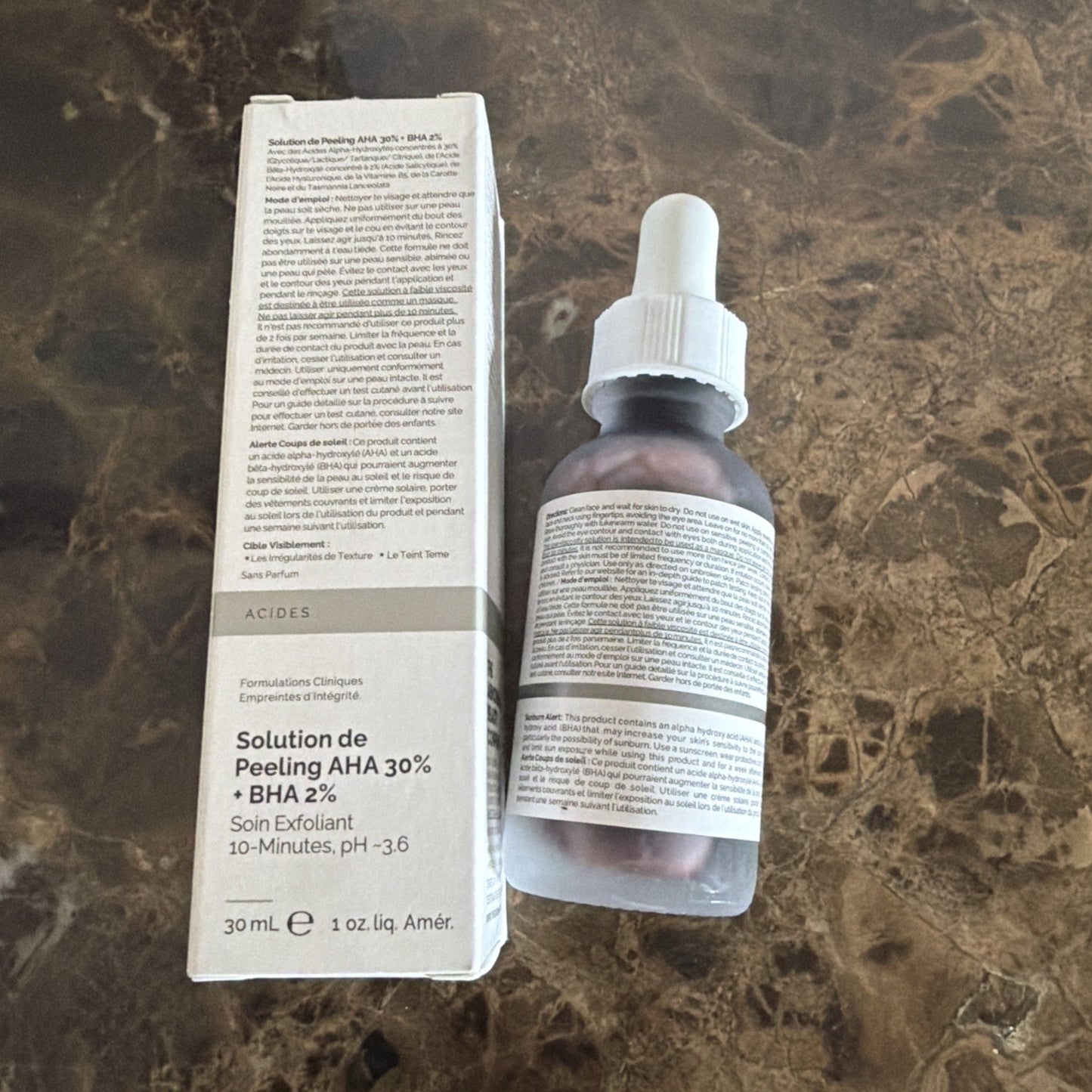 Small brown glass dropper bottle with a white label full of text next to a white rectangular box labeled in French for a 30% AHA and 2% BHA exfoliating peeling solution on a marble surface.