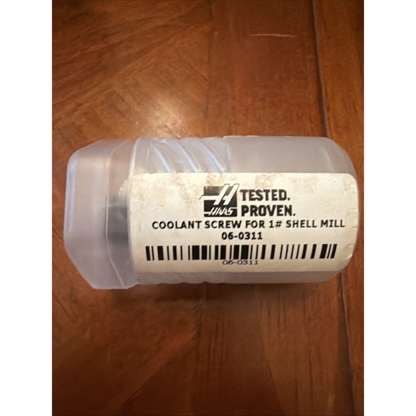 Small translucent plastic container with a white label showing the Haas logo, text indicating it holds a coolant screw for a 1-inch shell mill, and a barcode, placed on a wooden surface.