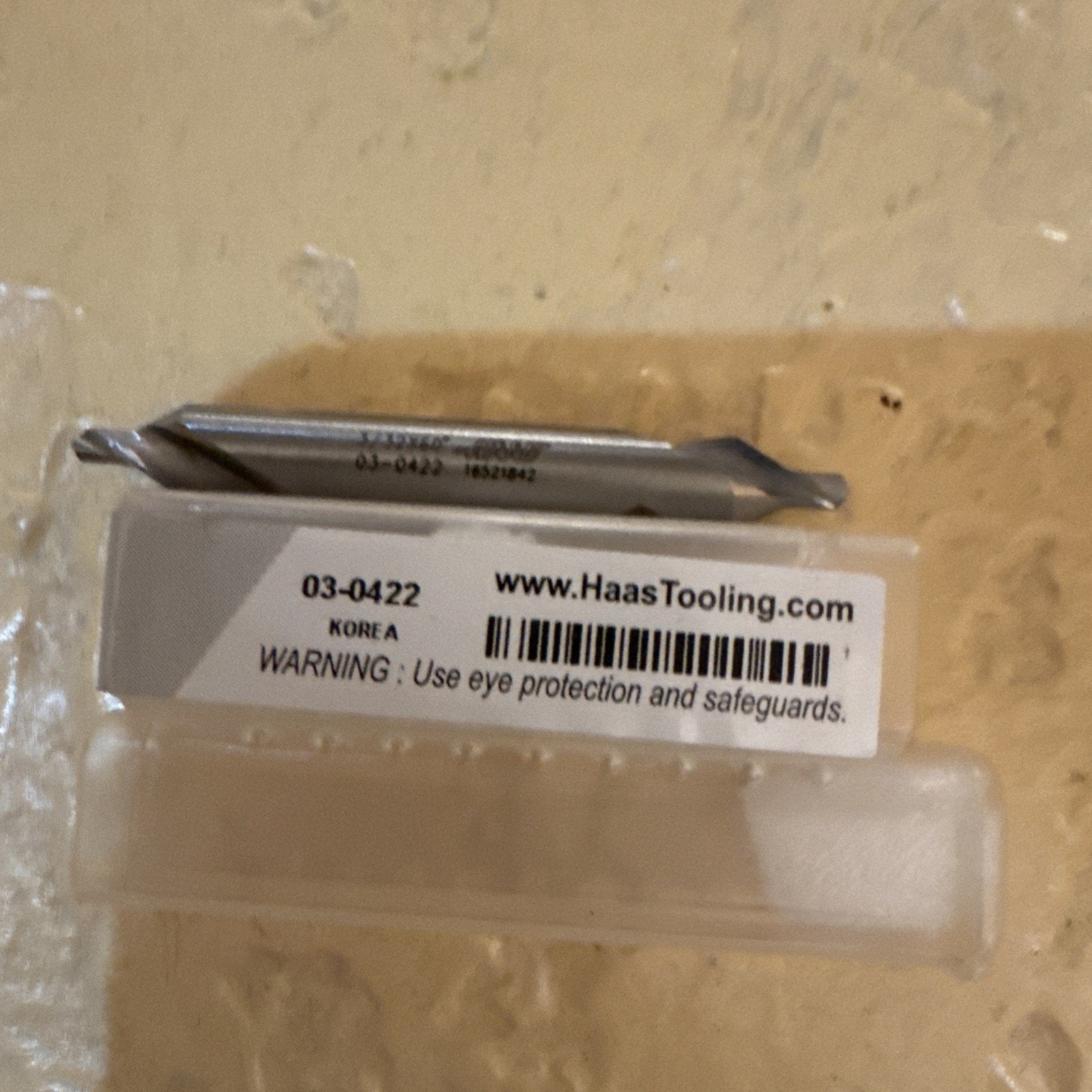 Close-up of a small metal cutting tool resting on top of a clear plastic case with a white label showing product code 03-0422, barcode, manufacturing origin Korea, and a safety warning to wear eye protection.