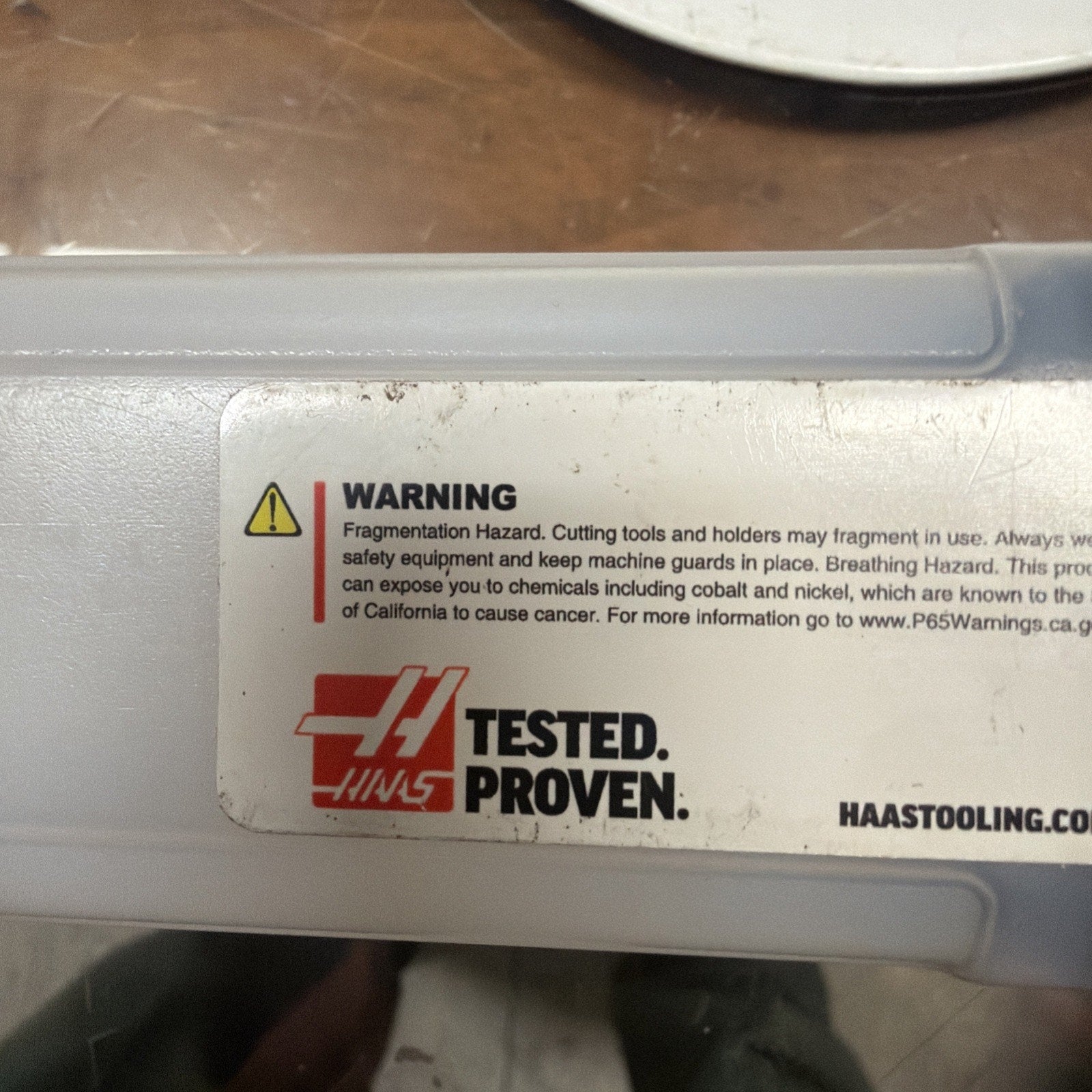 Close-up of a white plastic storage case with a warning label detailing fragmentation and breathing hazards, alongside a red and black logo and website URL, placed on a reflective surface.