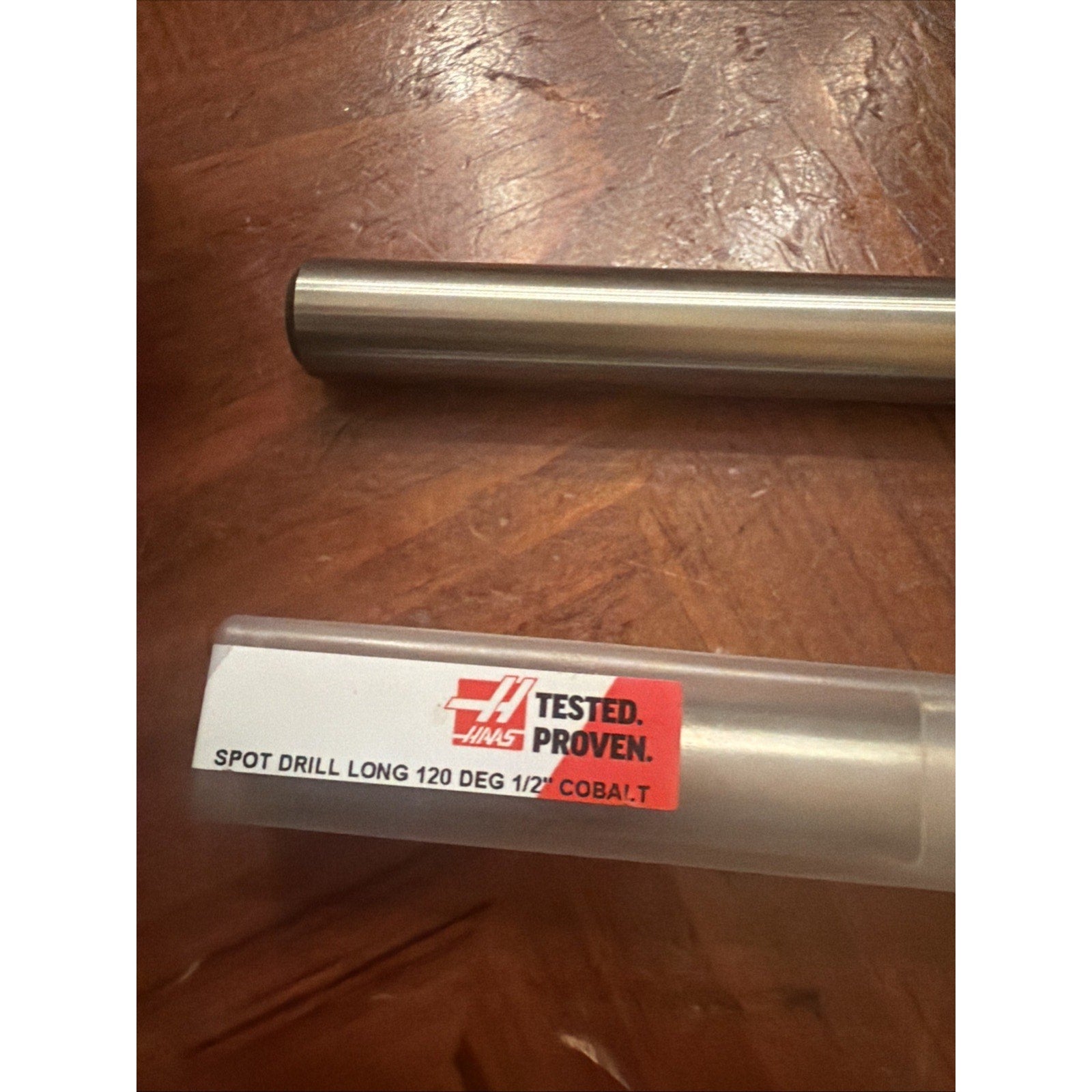 Metal drill bit resting beside a clear plastic case with a label reading "SPOT DRILL LONG 120 DEG 1/2" COBALT" and a red "TESTED. PROVEN." sticker, placed on a brown textured surface.