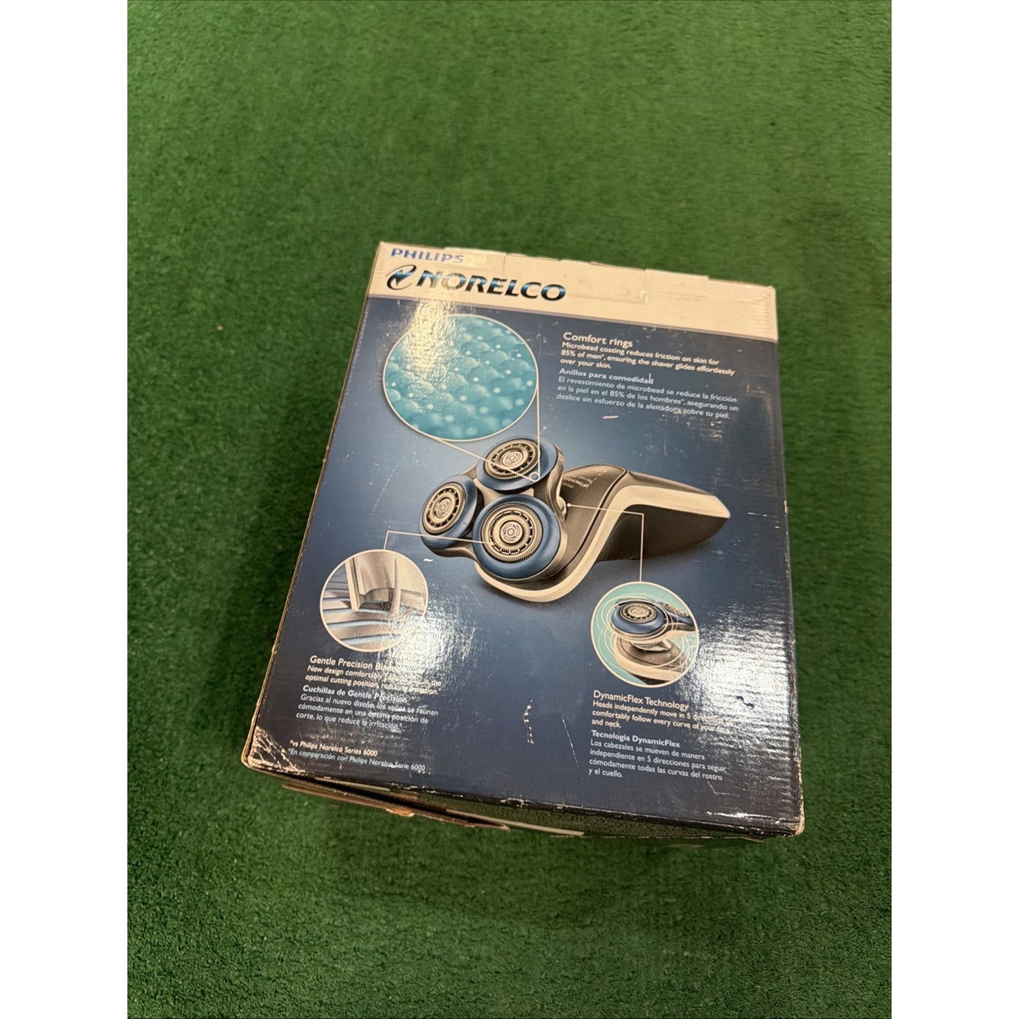 Worn retail box for a Philips Norelco electric shaver with images and descriptions highlighting comfort rings, gentle precision blades, and dynamic flex technology, placed on green carpet.