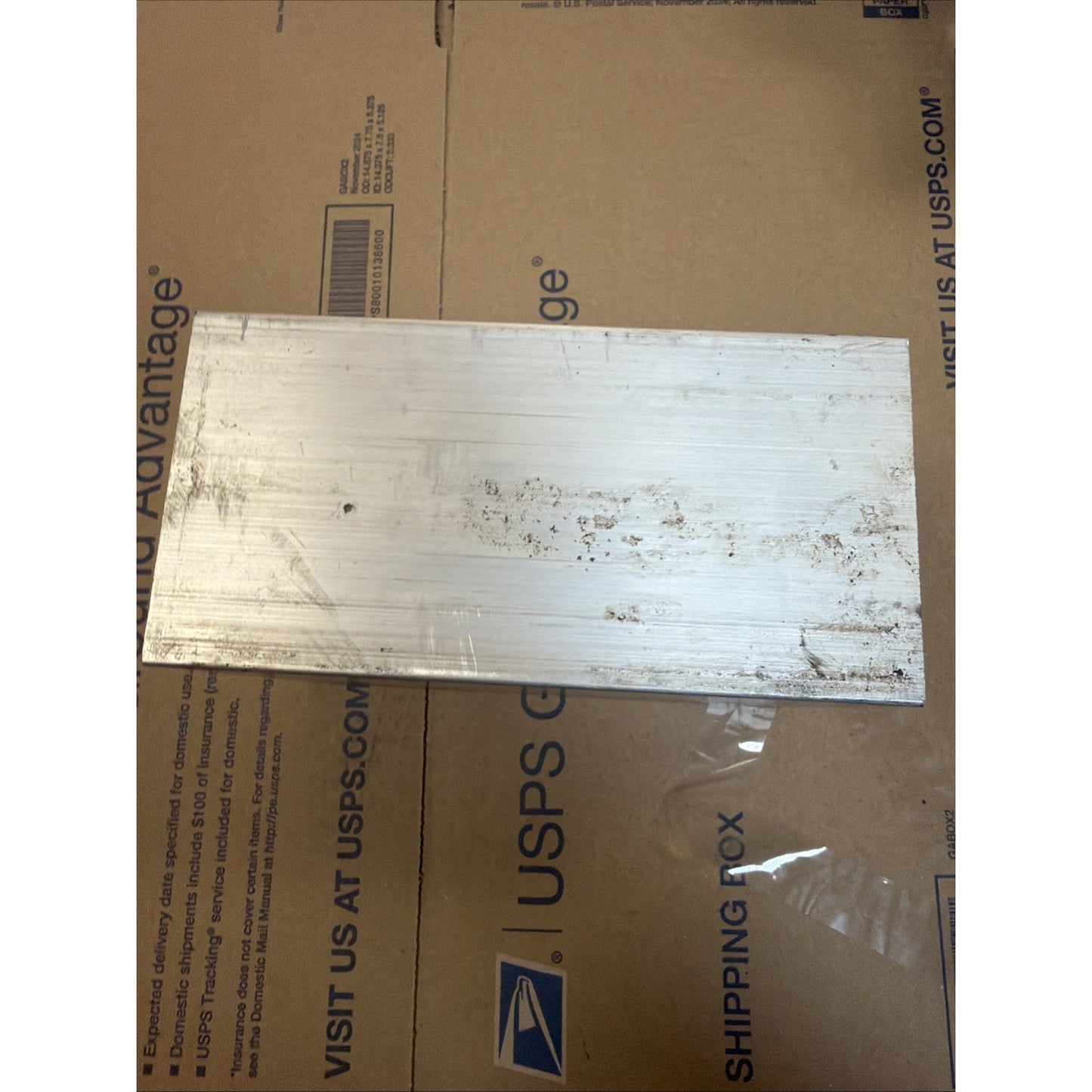 Rectangular aluminum metal plate with scratches and surface marks, placed on a USPS shipping box with visible text and logos.