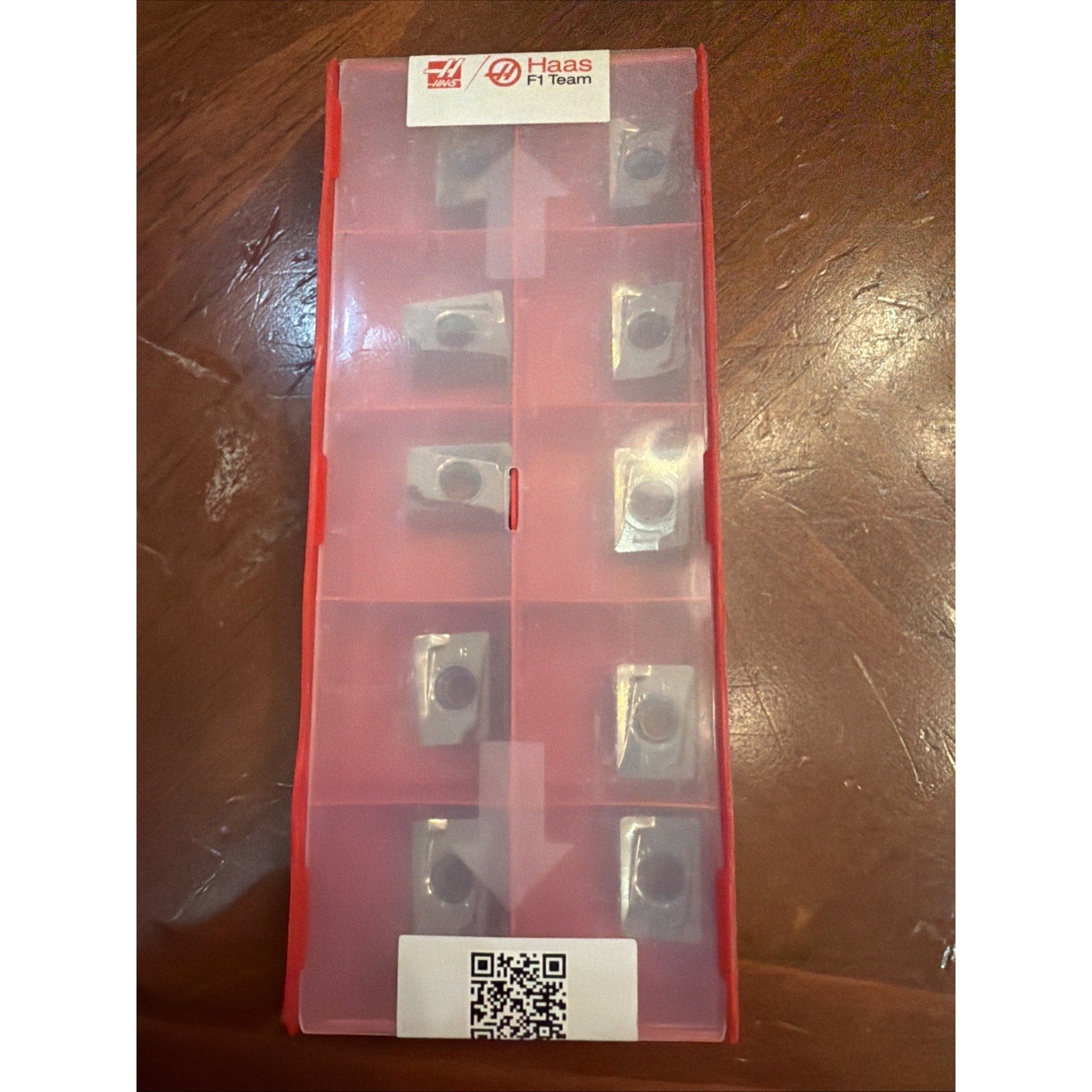 Red translucent plastic storage box with ten metal cutting inserts arranged in separate compartments, featuring a Haas F1 Team label and a QR code, placed on a wooden surface.