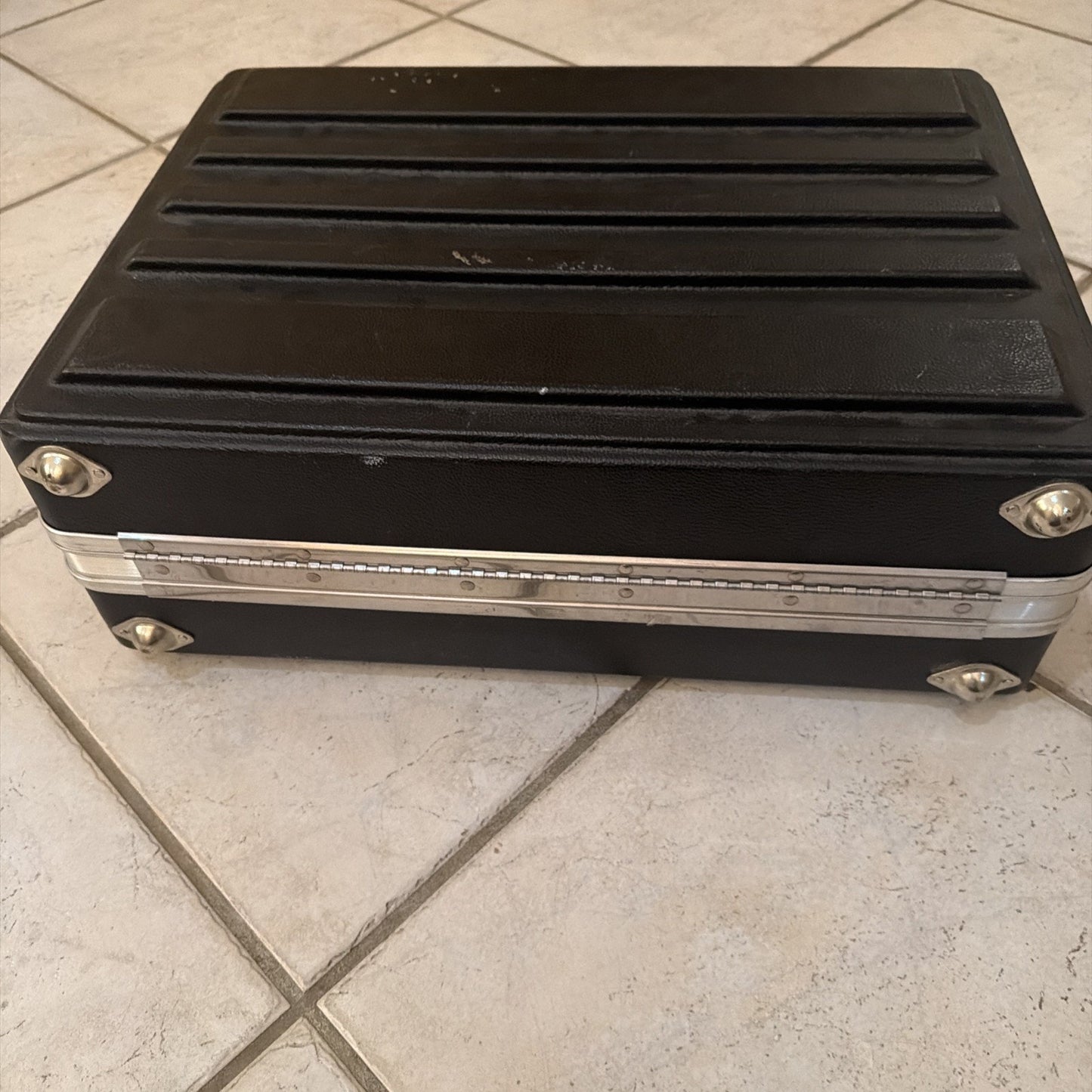 Rectangular black hard plastic carrying case with horizontal ridges on top, silver metal corner protectors and a latch, placed on a light-colored tiled floor.