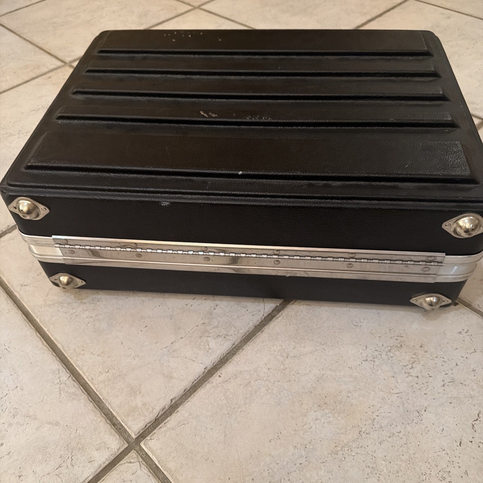 Rectangular black hard plastic carrying case with horizontal ridges on top, silver metal corner protectors and a latch, placed on a light-colored tiled floor.