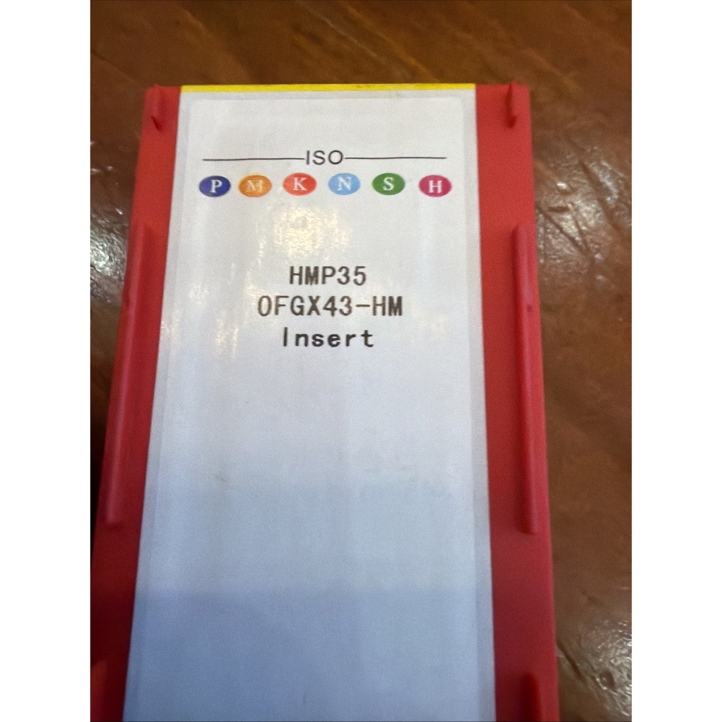 Red plastic storage container with a white label displaying ISO classification symbols, product code "HMP35 0FGX43-HM," and the word "Insert" on a wooden surface.