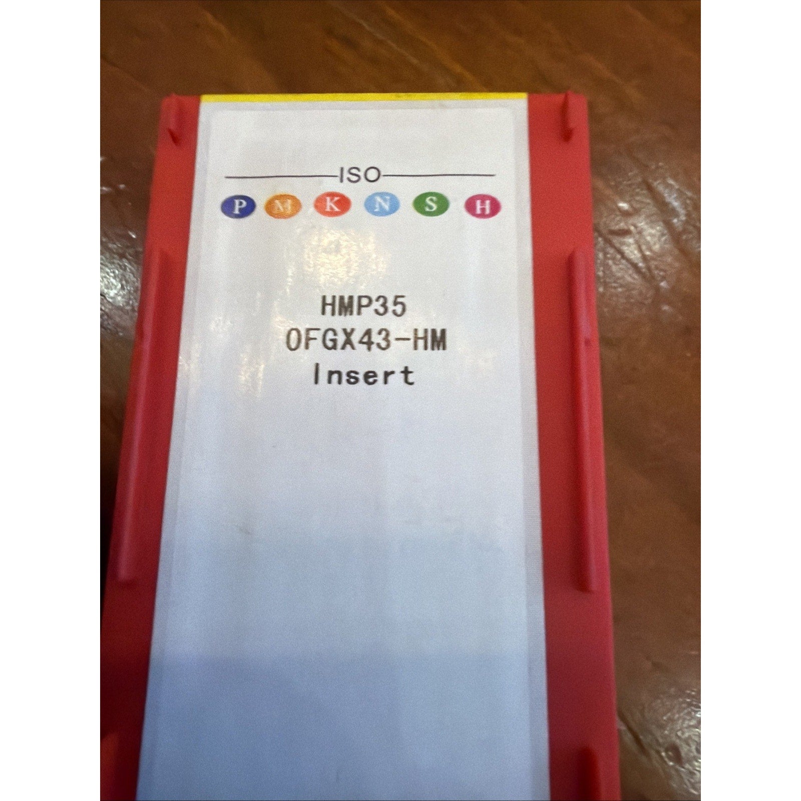 Red plastic storage container with a white label displaying ISO classification symbols, product code "HMP35 0FGX43-HM," and the word "Insert" on a wooden surface.