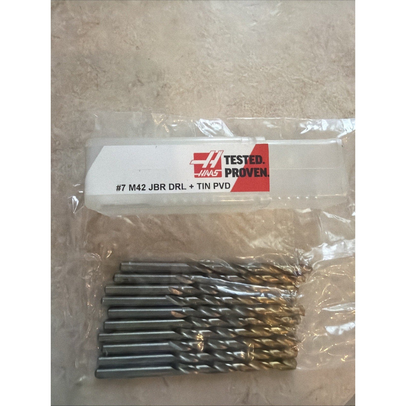 Set of ten metal drill bits with spiral flutes arranged side by side inside a clear plastic package labeled with red and black text indicating "#7 M42 JBR DRL + TIN PVD" on a beige textured surface.
