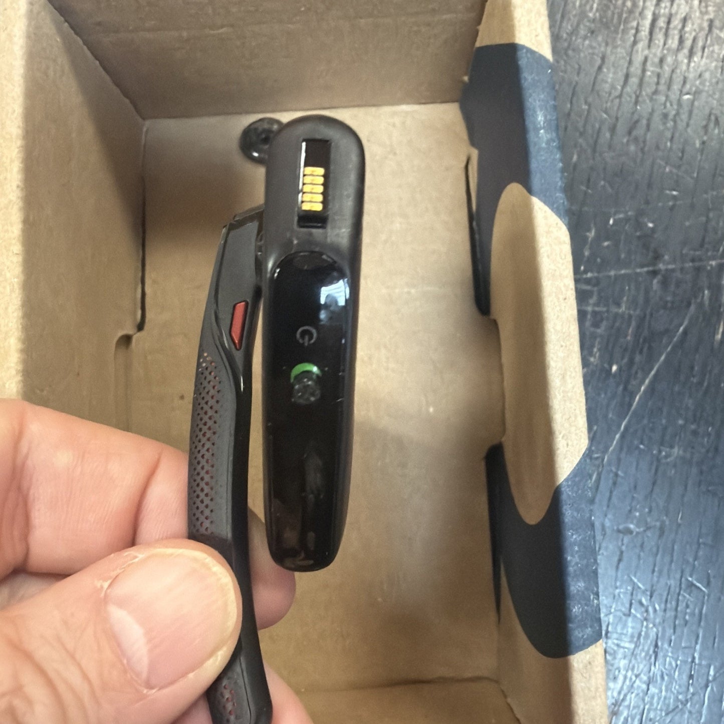 Hand holding a black fitness tracker with a digital display and charging pins, shown inside a partially open cardboard box on a dark wooden surface.