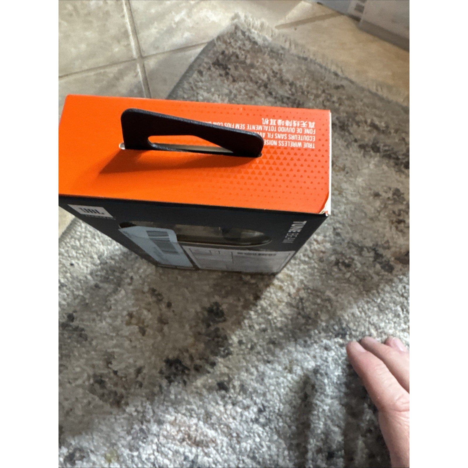 JBL by Harman Tune Beam Earbuds black box with orange top and black handle placed on carpet near hand