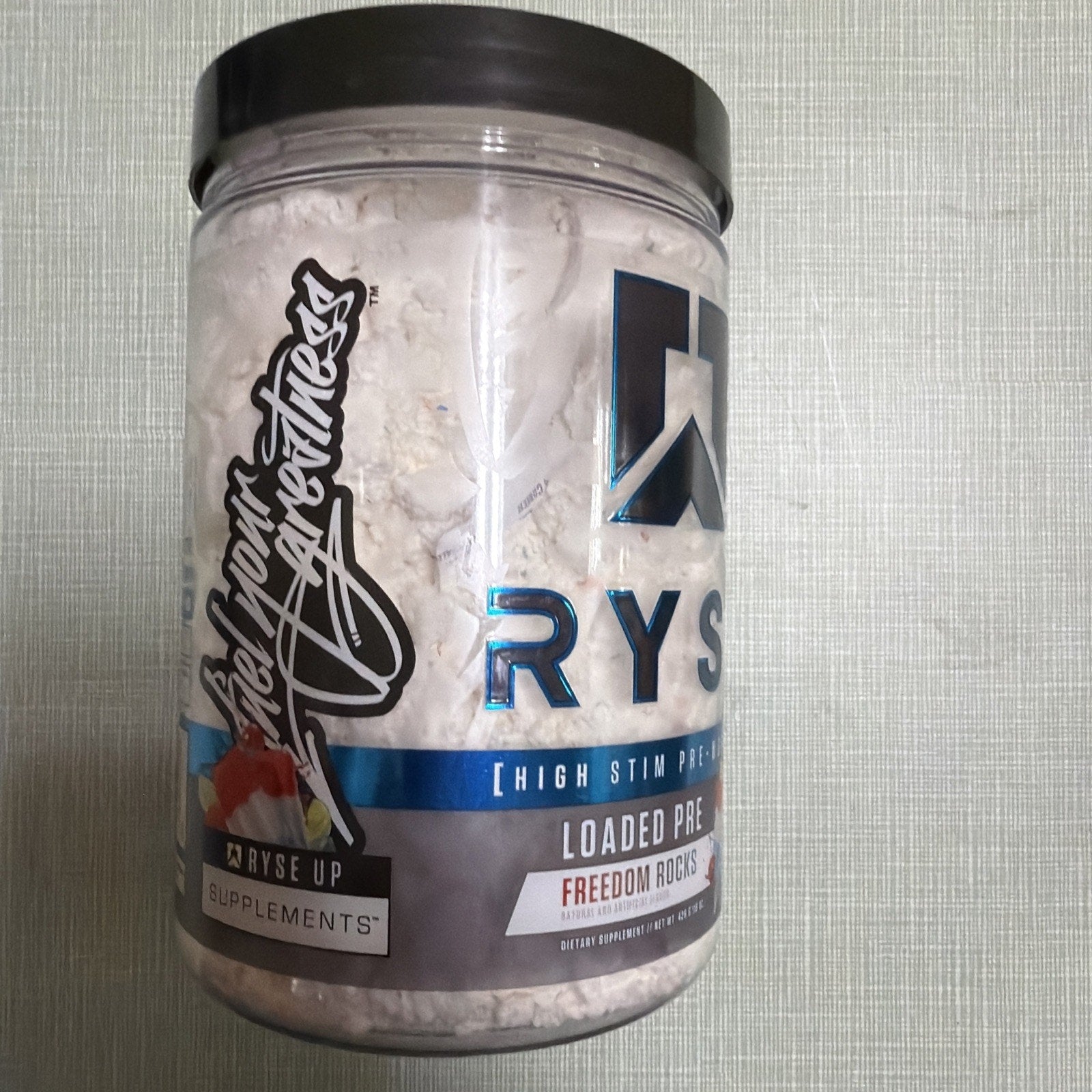 Clear plastic container with white powder supplement labeled "Loaded Pre" by Ryse Up Supplements, featuring a black lid and a design element resembling red and green fruit on a gray background.