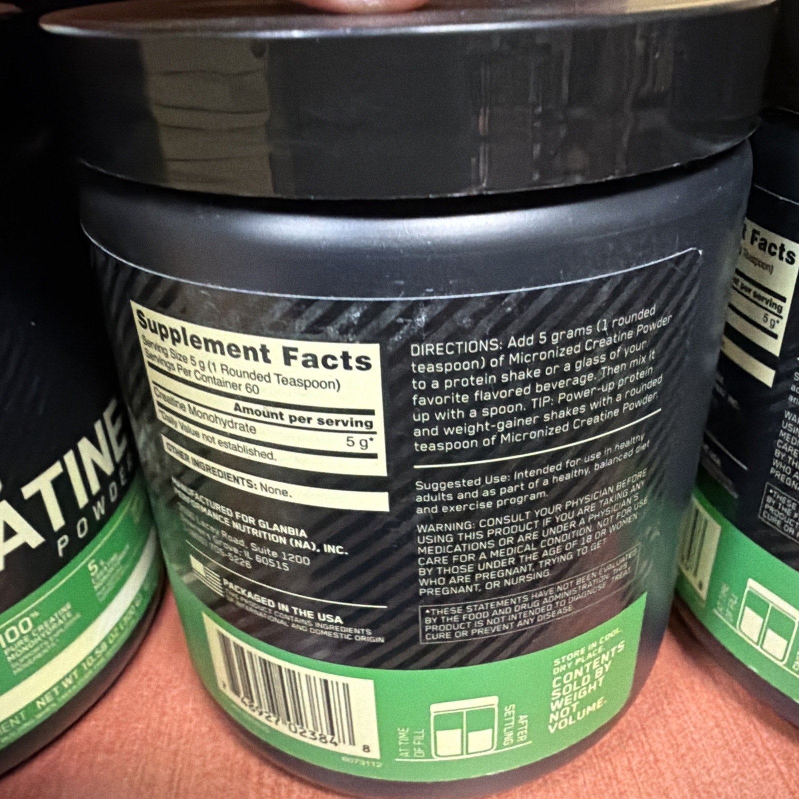 Close-up of a black container label showing supplement facts for creatine monohydrate powder with 5 grams per serving and directions to mix one rounded teaspoon with water or a protein shake.