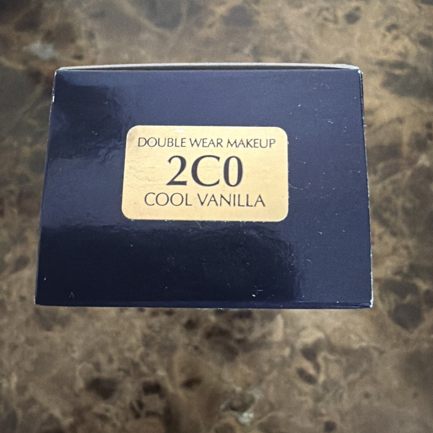 Top view of a dark blue rectangular box with a beige label displaying "Double Wear Makeup 2C0 Cool Vanilla," placed on a brown marbled surface.