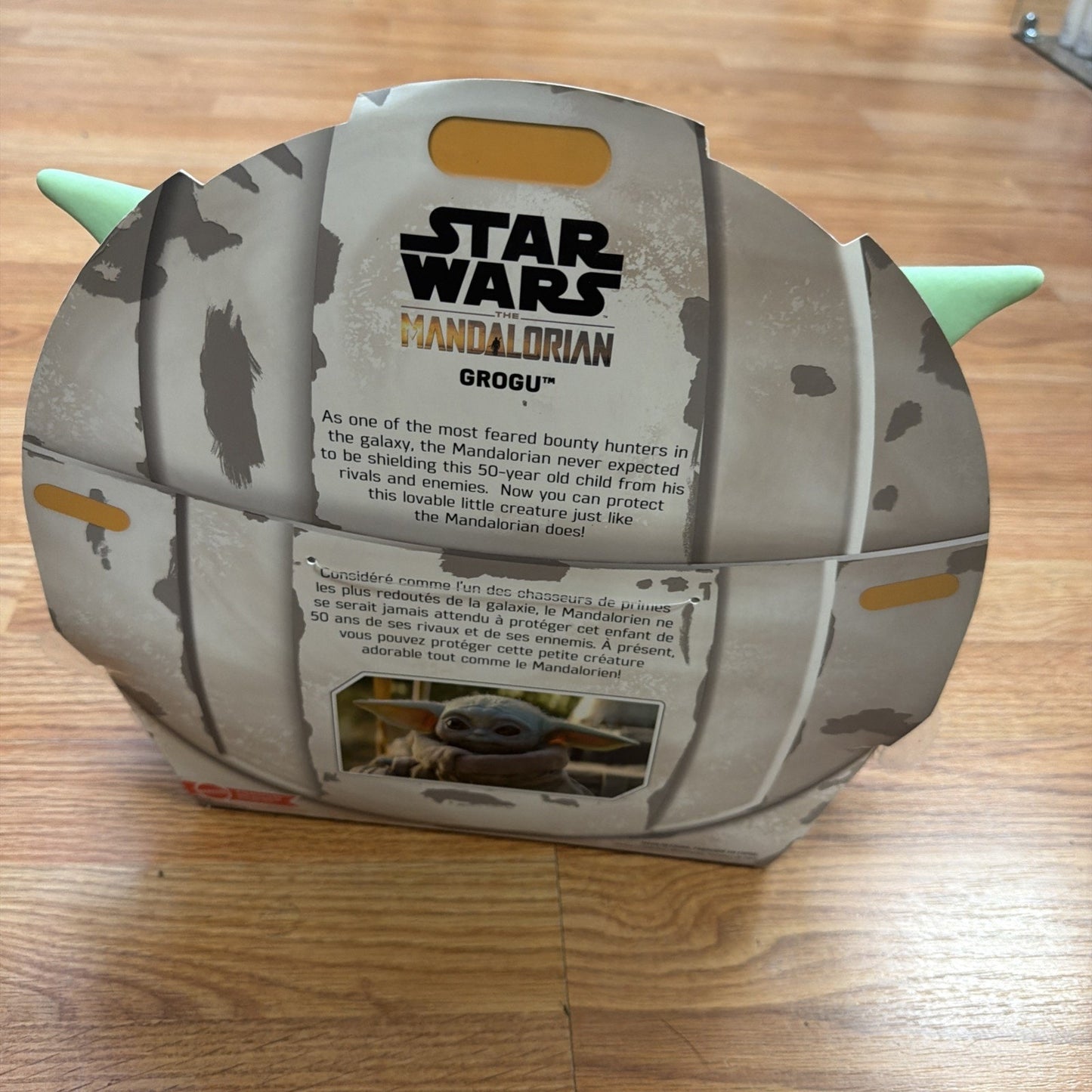 Cardboard packaging shaped like Baby Yoda's head from Star Wars The Mandalorian, featuring text about Grogu and a small image of the character on a wooden floor background.