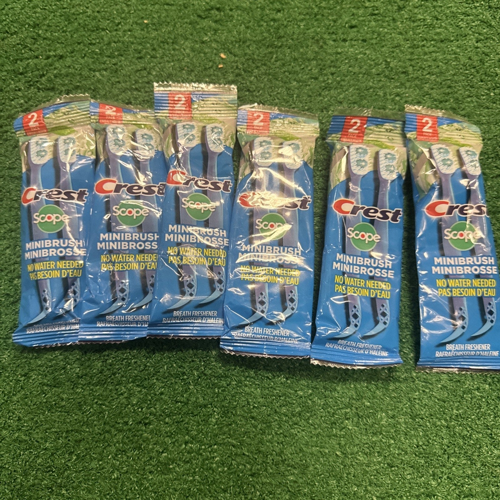 Six packs of Crest Scope waterless disposable minibrushes with toothpaste and pick shown on grass background