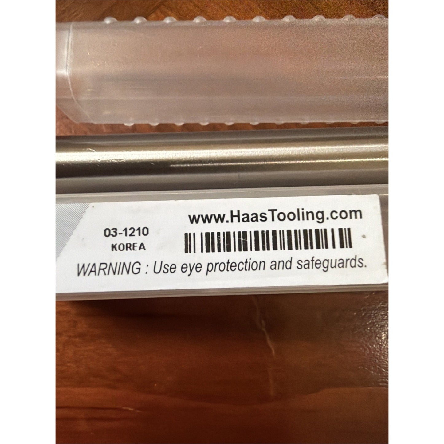 Close-up of a white label on a metal tool with the website HaasTooling.com, a barcode, product code 03-1210, "KOREA" printed underneath, and a warning to use eye protection and safeguards, resting on a wooden surface with a clear plastic tube above.