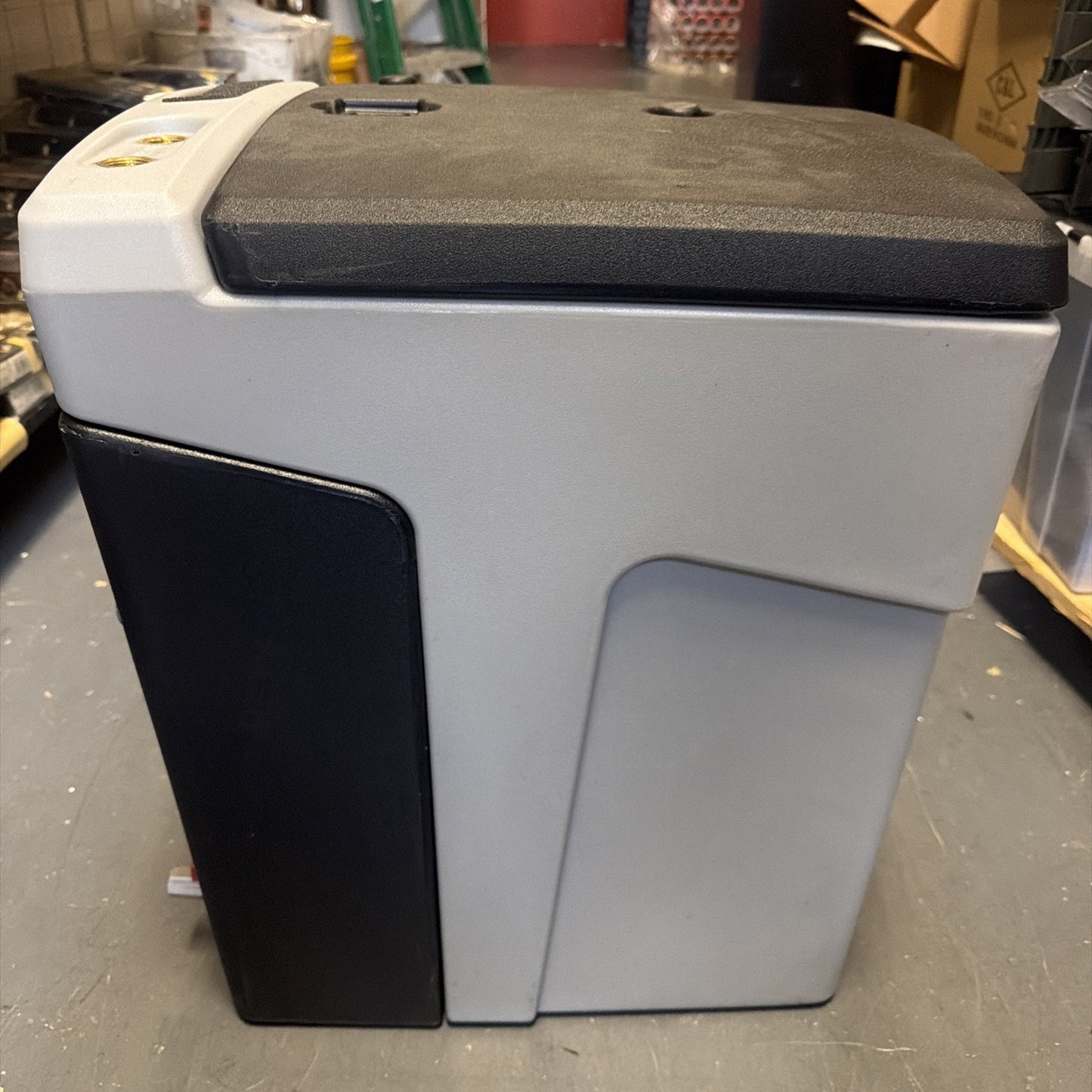 Side view of a compact gray and black electronic device with a textured top surface, placed on a gray floor in an industrial or storage setting.